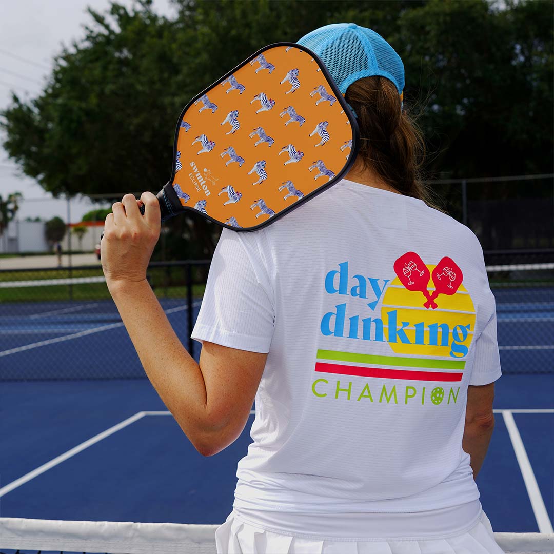 A woman stands on a blue tennis court holding a Swinton Pickleball Eclipse Party Animal paddle with an orange, textured graphite face. She wears a blue cap and a “day drinking champion” shirt with colorful graphics and red paddles.