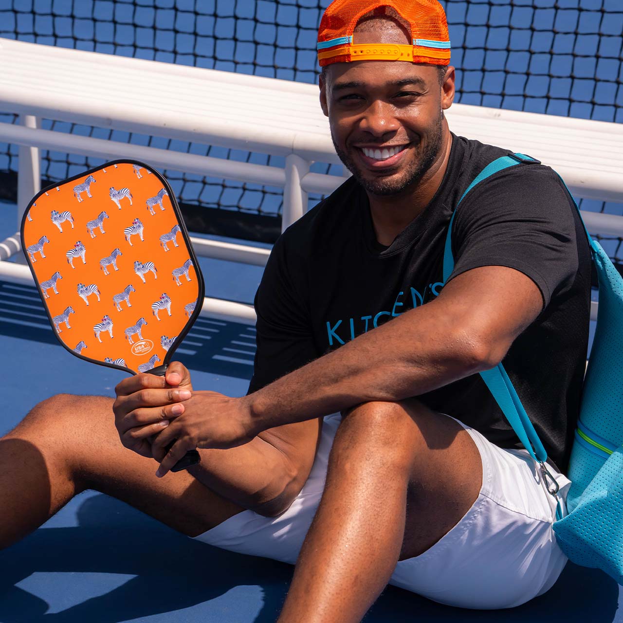 A smiling man in athletic wear sits on a blue court holding the Swinton Pickleball Eclipse Party Animal Paddle with a blue and white dog pattern. He wears a backwards orange cap, black shirt, and blue backpack; a net and bench are visible behind him.