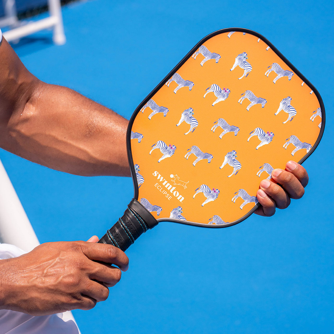 A player grips the Swinton Pickleball Eclipse Party Animal Paddle, featuring an orange graphite face with cartoon zebras and a black handle, standing out vividly against a blue court. USA Pickleball Approved.