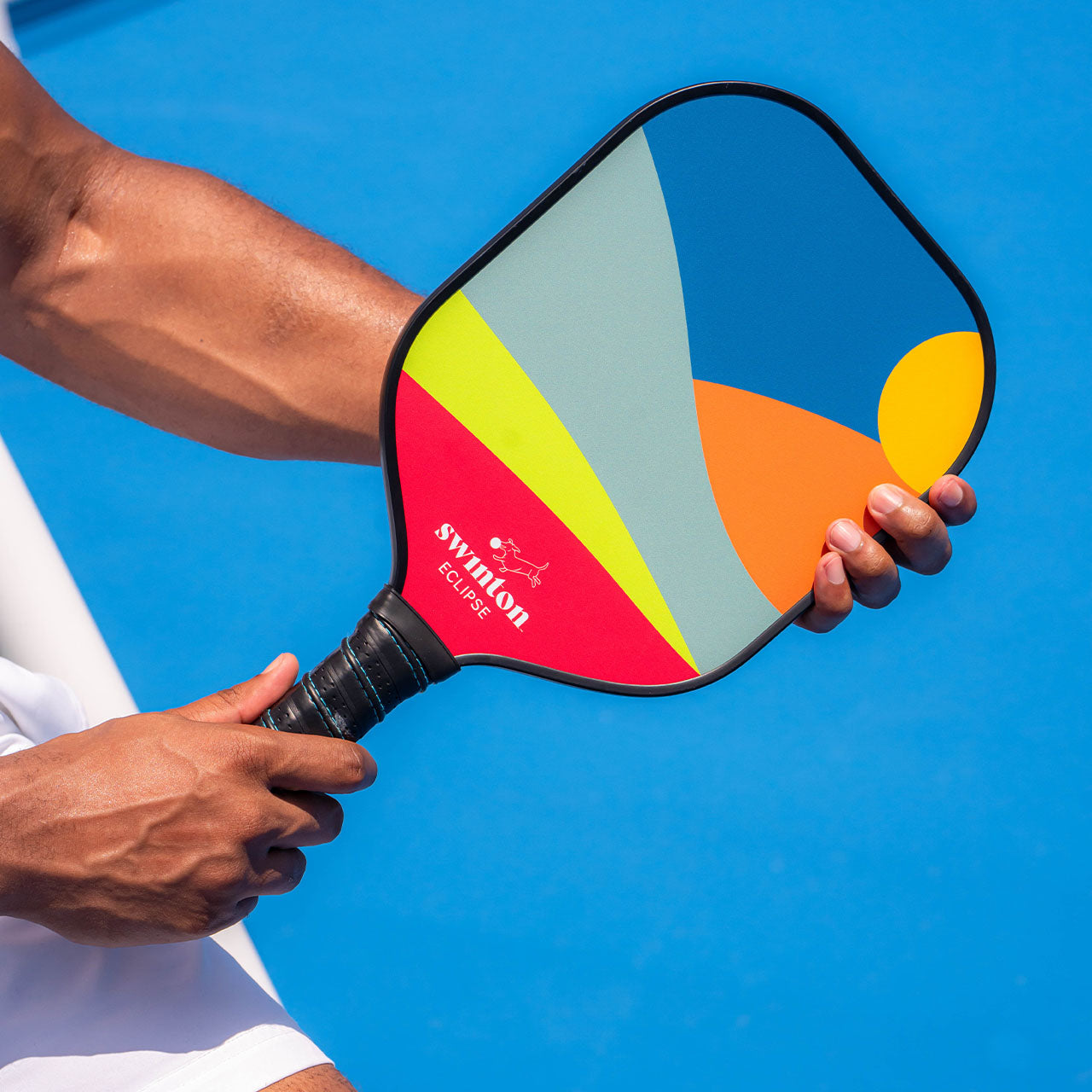 A person holds the Swinton Pickleball Eclipse Sunset paddle, USA Pickleball Approved, featuring a modern abstract design in blue, green, yellow, orange, and gray against a bright blue court and white clothing.
