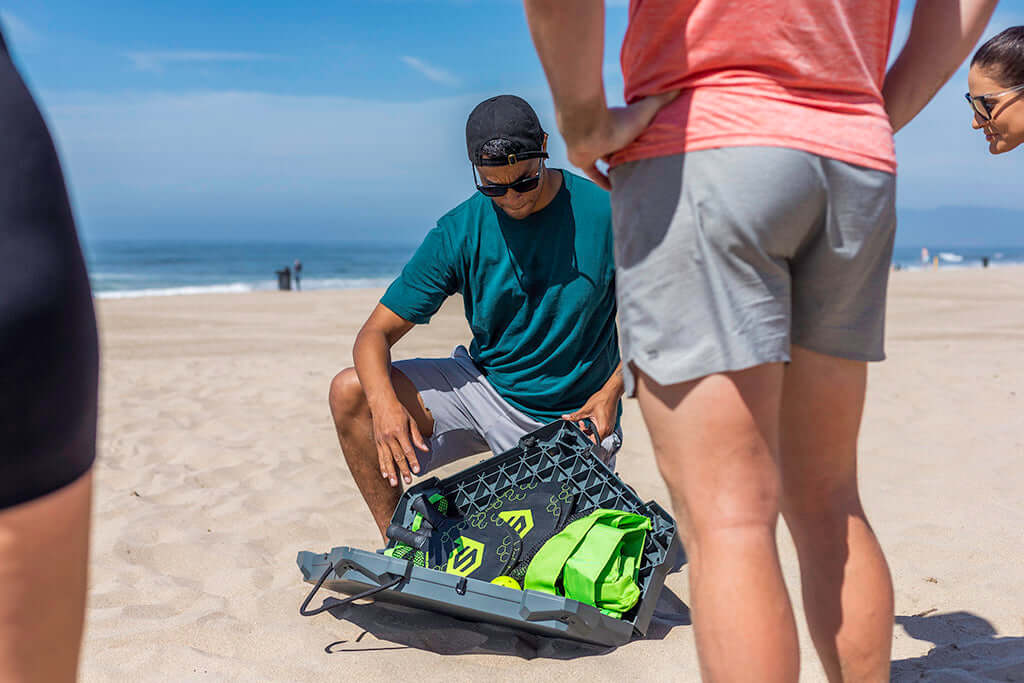 A man in sunglasses and a backwards cap kneels on the beach, packing neon green PaddleSmash game items into a foldable tote. Two others stand nearby, with the ocean and blue sky visible in the background. Product: PaddleSmash by PaddleSmash.