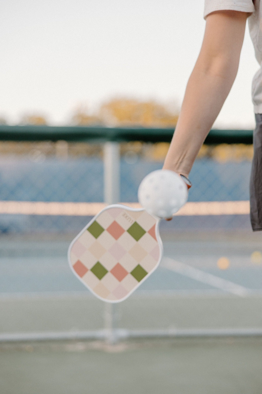 A person swings the LUXE Pickleball Party Checker paddle—pastel check-patterned, USA Pickleball Approved—hitting a white ball on a blue outdoor court with a blurred green fence and scattered yellow balls in the background.