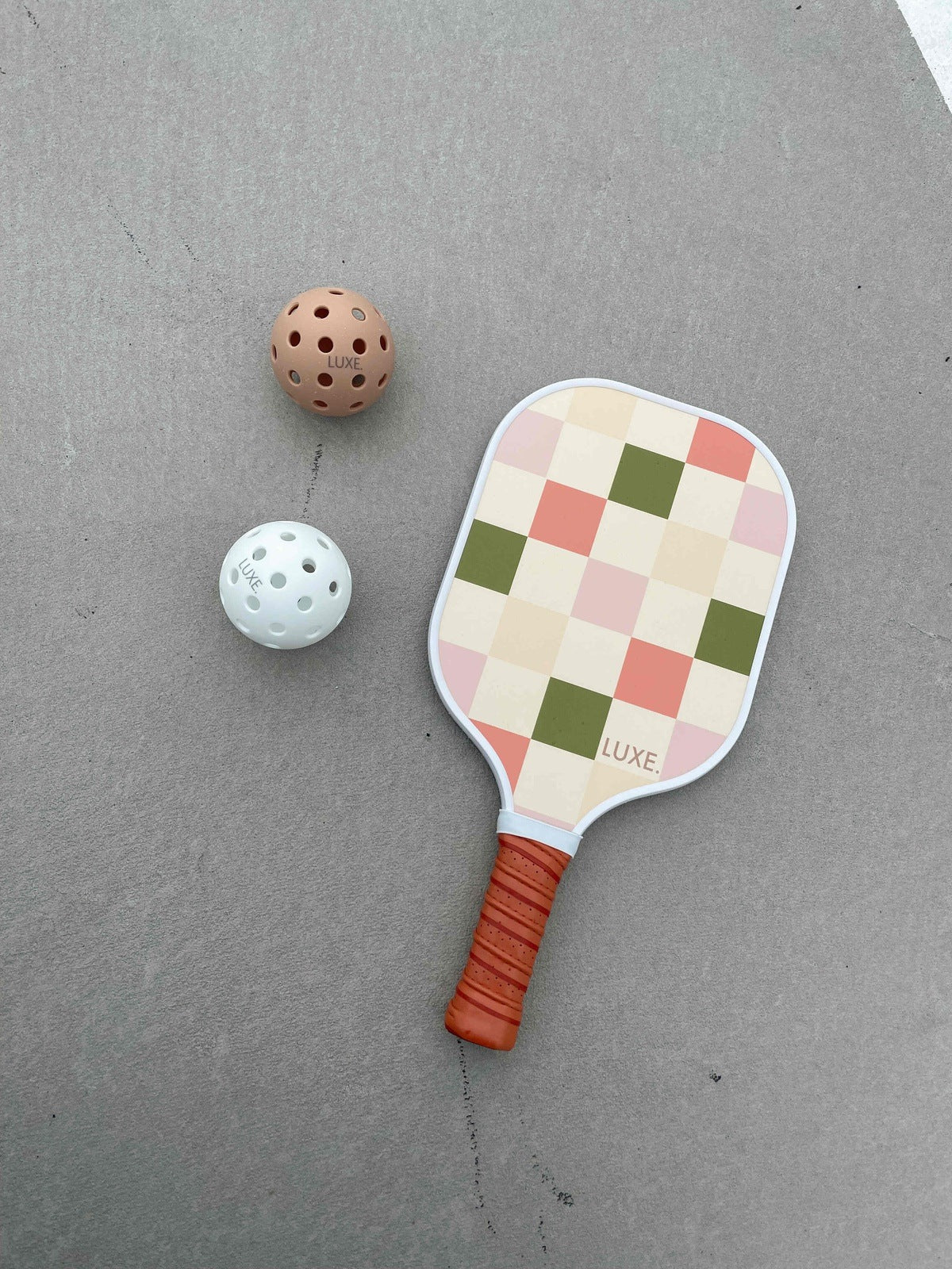 The LUXE Pickleball Party Checker paddle, featuring a pink, green, and cream checkered design with an orange handle, sits on a gray surface beside two pickleballs. The paddle is USA Pickleball Approved.