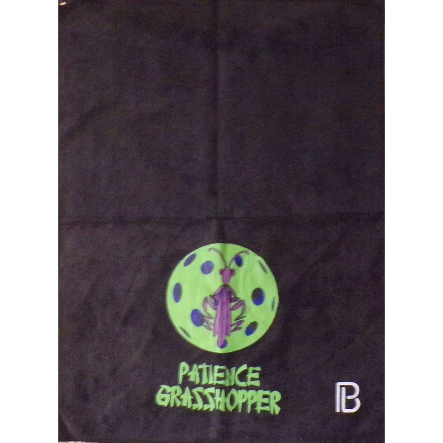 The Pickleball Bella Patience Pickleball Towel displays a green circle with a purple grasshopper and dark spots on black polyester, "PATIENCE GRASSHOPPER" in green text, and a white-and-black PB logo in the bottom right corner.