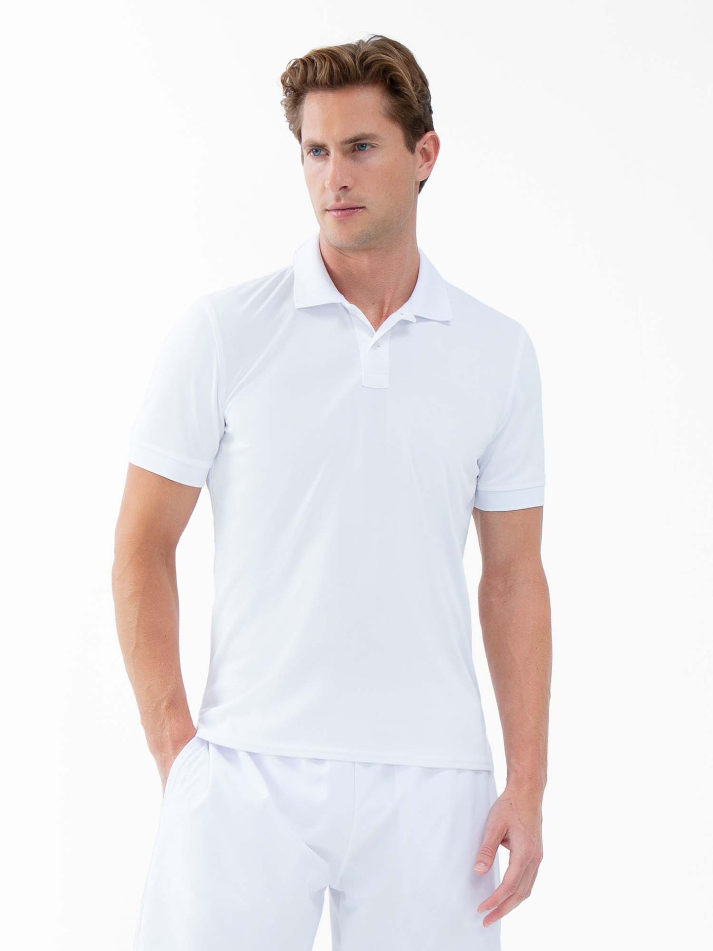 A man with short, light brown hair stands against a white backdrop in the inPhorm NYC Performance Men's Classic Tennis Rib Collar Polo - White and white pants, showcasing its UV protection and moisture-wicking features with a relaxed, neutral pose.