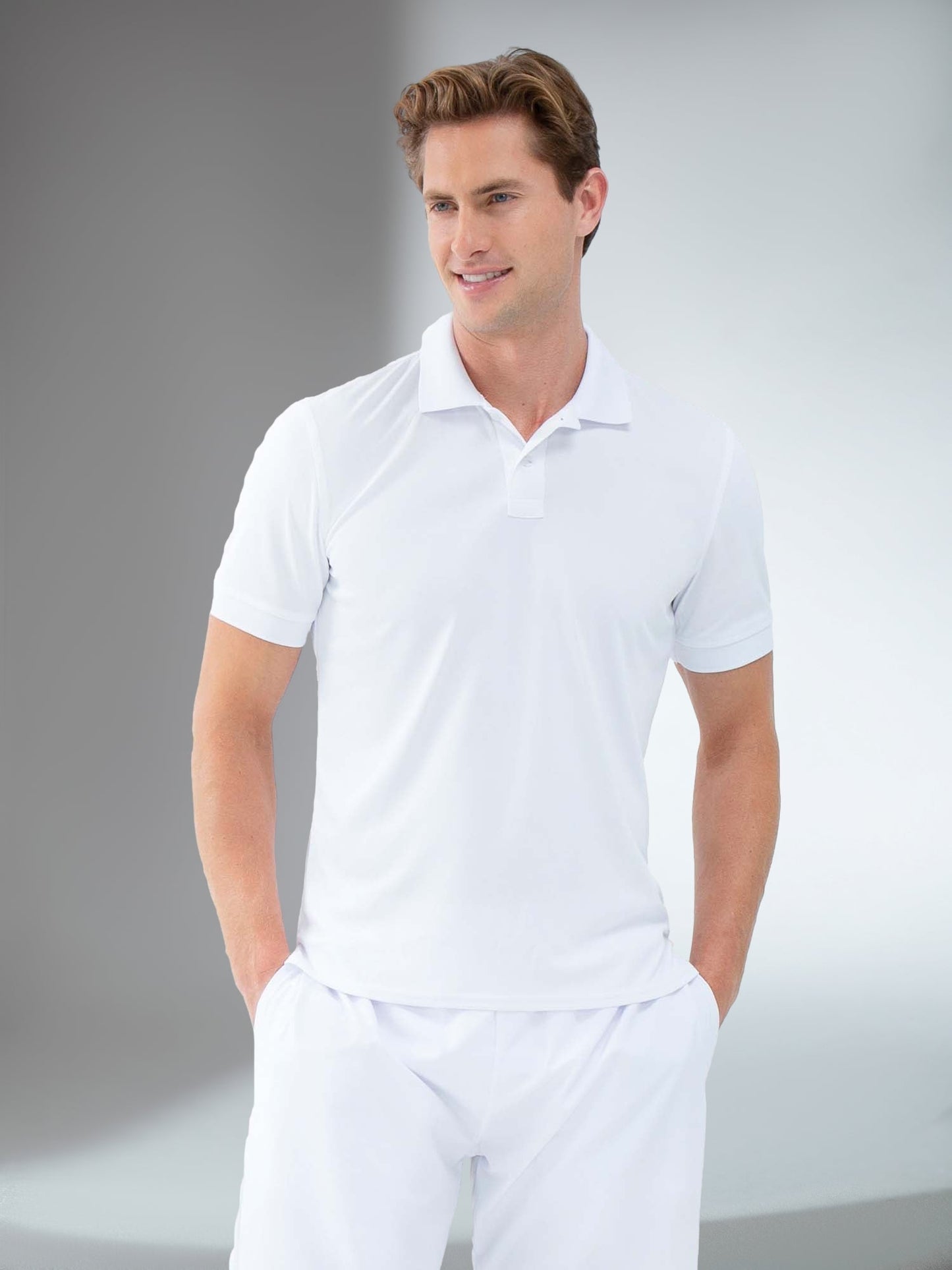 A man with short, light brown hair wears the inPhorm NYC Performance Men's Classic Tennis Rib Collar Polo in white and white pants, smiling against a gray and white background as he enjoys the polo's moisture-wicking comfort.
