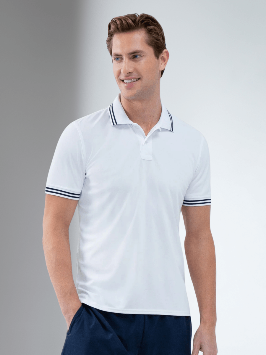 A smiling man with short, light brown hair wears the inPhorm NYC Performance Men's Classic Tennis Rib Collar Polo in White/Midnight. The polo features moisture-wicking fabric and dark striped trim. He stands against a neutral background, hand in his navy pants pocket.
