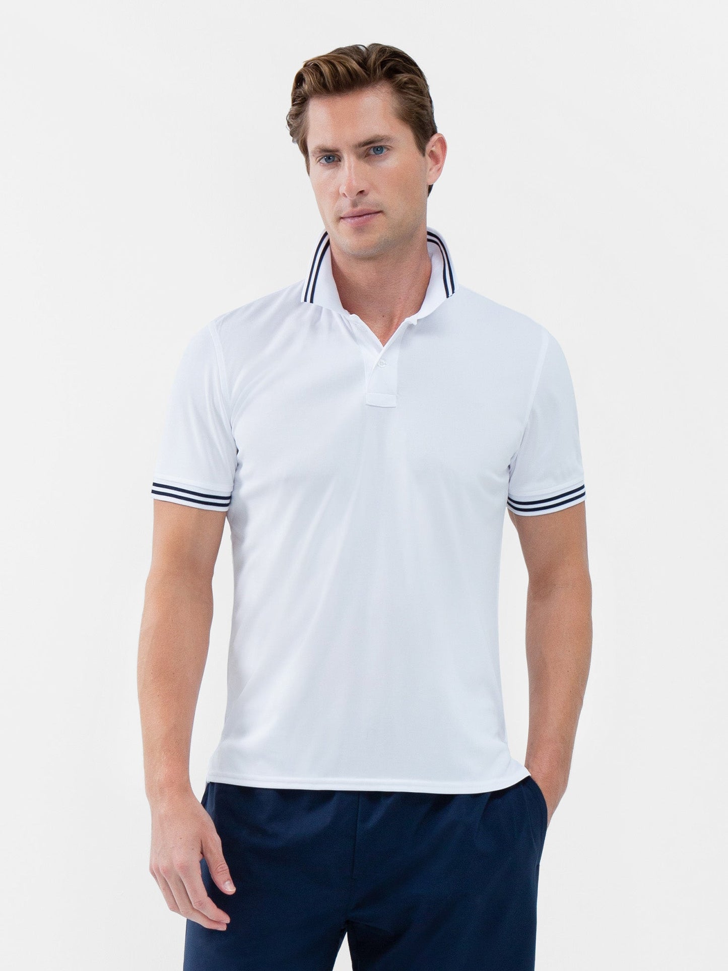 A man with light brown hair wears the inPhorm NYC Performance Men's Classic Tennis Rib Collar Polo in White/Midnight, featuring navy stripes on the collar and cuffs, paired with navy pants. He stands facing forward against a plain white background.