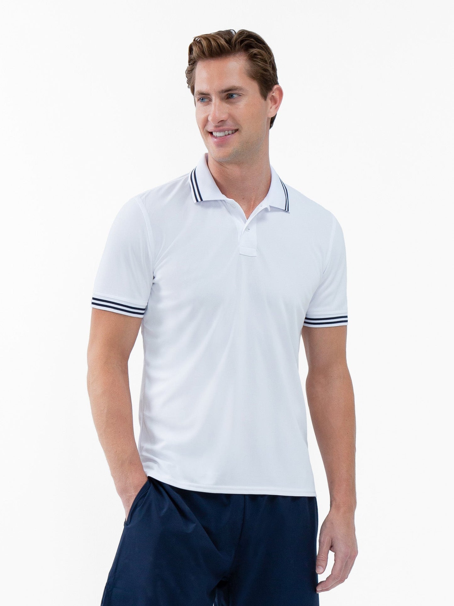 A man with light skin and short brown hair smiles while wearing the inPhorm NYC Performance Men's Classic Tennis Rib Collar Polo in White/Midnight, paired with dark navy shorts, standing against a plain white background.