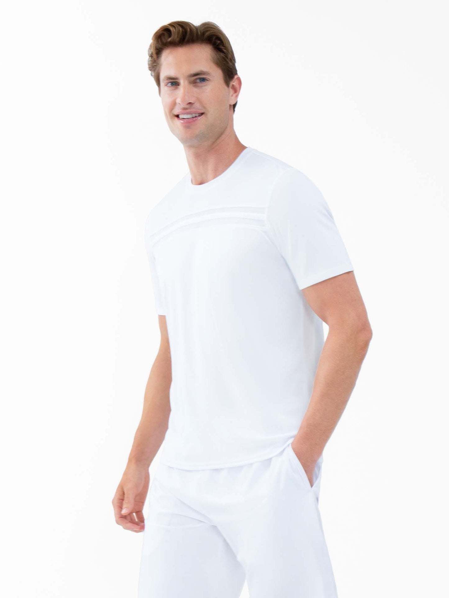 A man with light brown hair smiles at an angle, wearing the inPhorm NYC Performance Men’s Tournament Short Sleeve Crew Neck in white and matching pants. His left hand is in his pocket, set against a white background for a clean, minimal look.