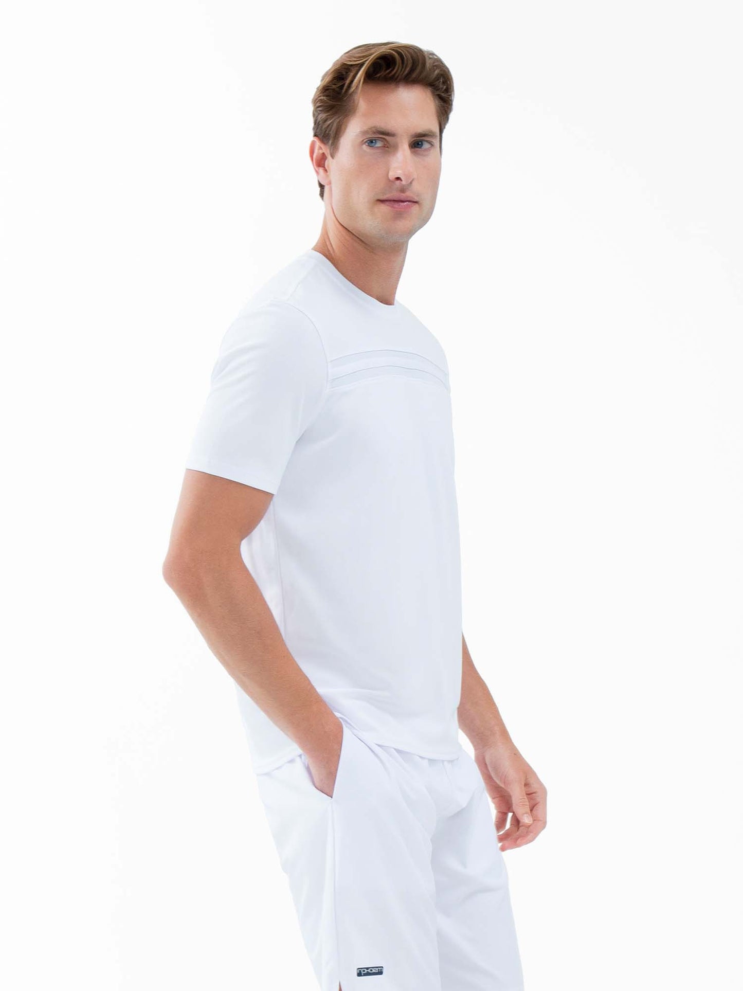 A man with short light brown hair wears the inPhorm NYC Performance Men's Tournament Short Sleeve Crew Neck in white, featuring subtle horizontal detail. He pairs it with matching pants and stands angled to the side against a plain white background.