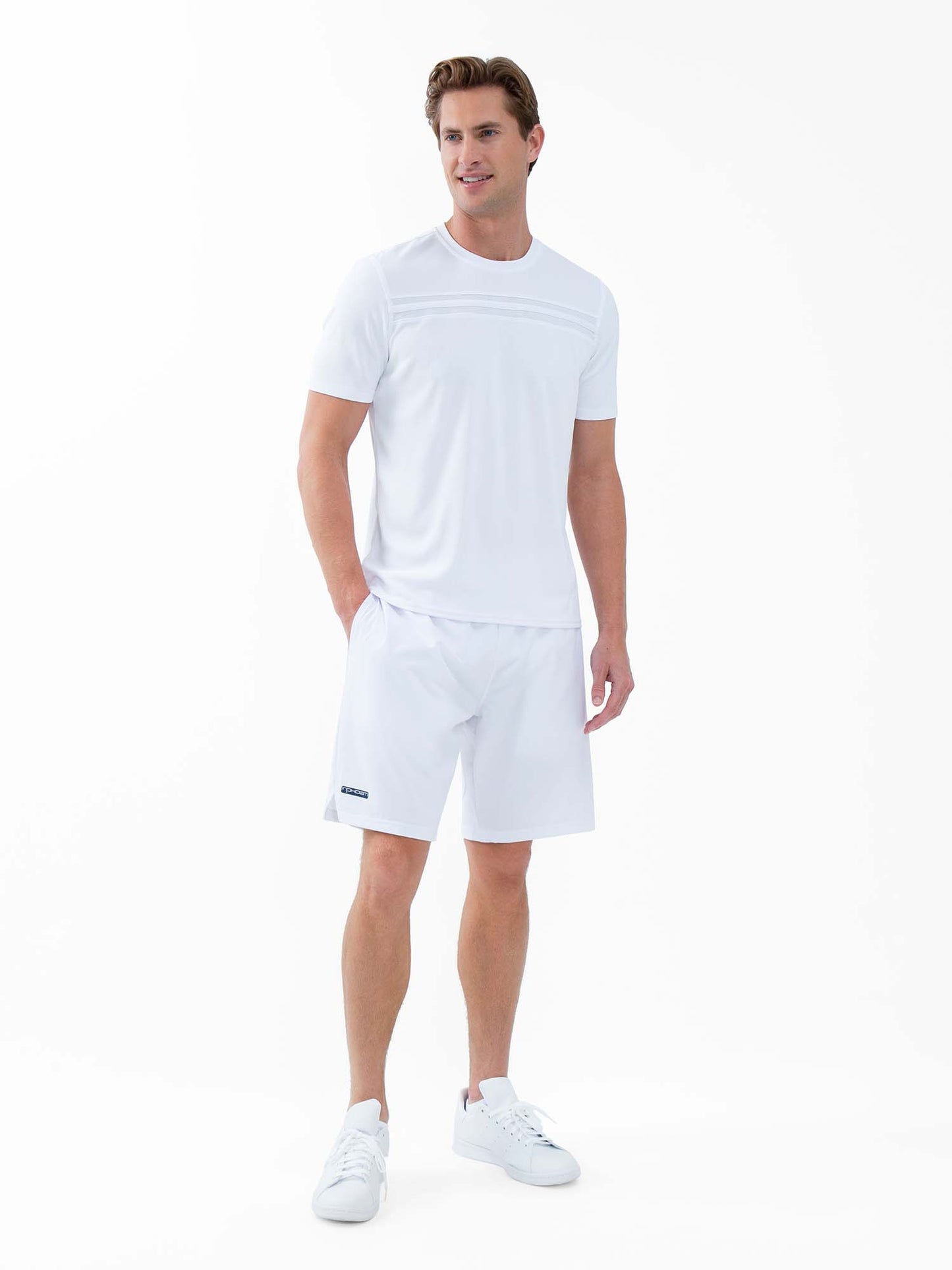 A man in a plain white background wears the inPhorm NYC Performance Men's Tournament Short Sleeve Crew Neck - White, paired with white shorts and sneakers. He smiles slightly, one hand in his pocket, with neatly styled short brown hair.
