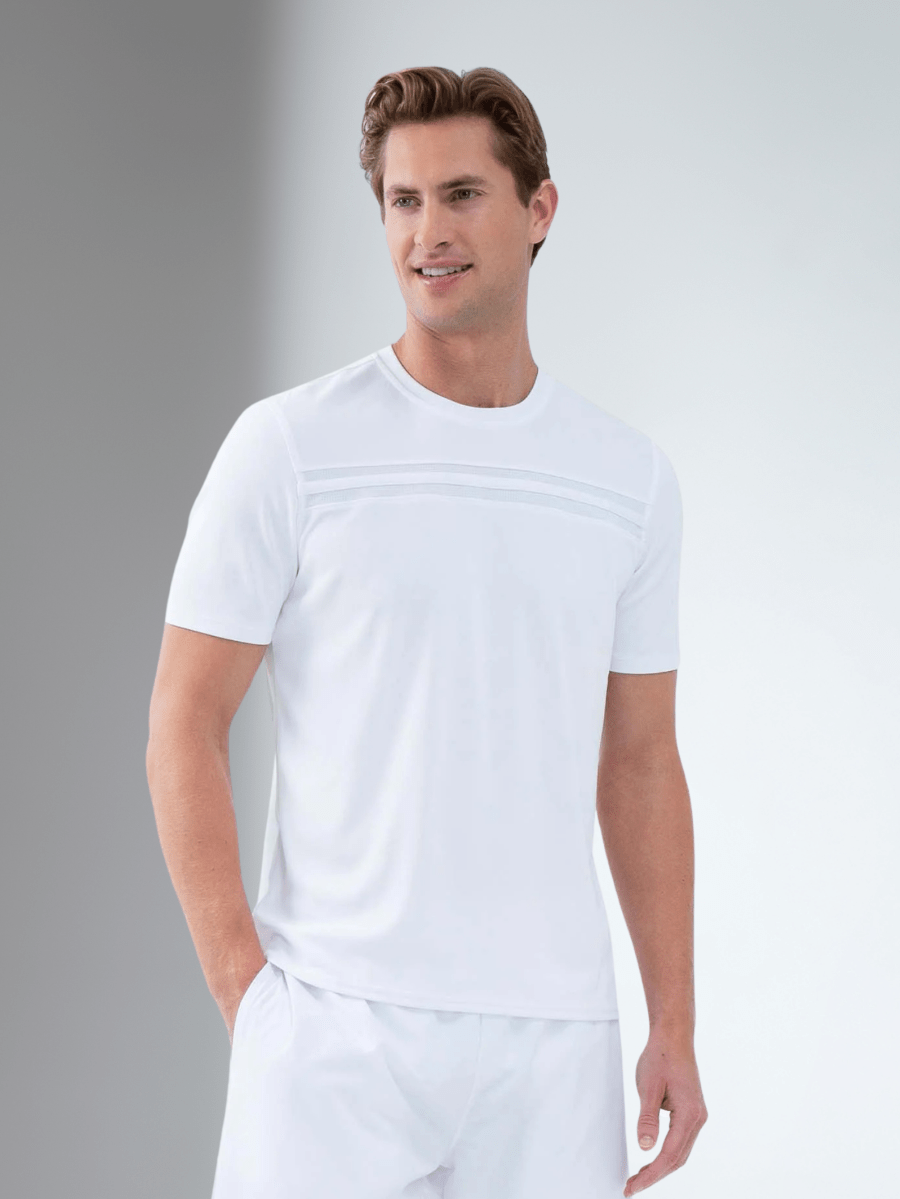 A man with light brown hair models the inPhorm NYC Performance Men's Tournament Short Sleeve Crew Neck in white, paired with white shorts, standing against a gray and white backdrop and smiling while looking to his left.