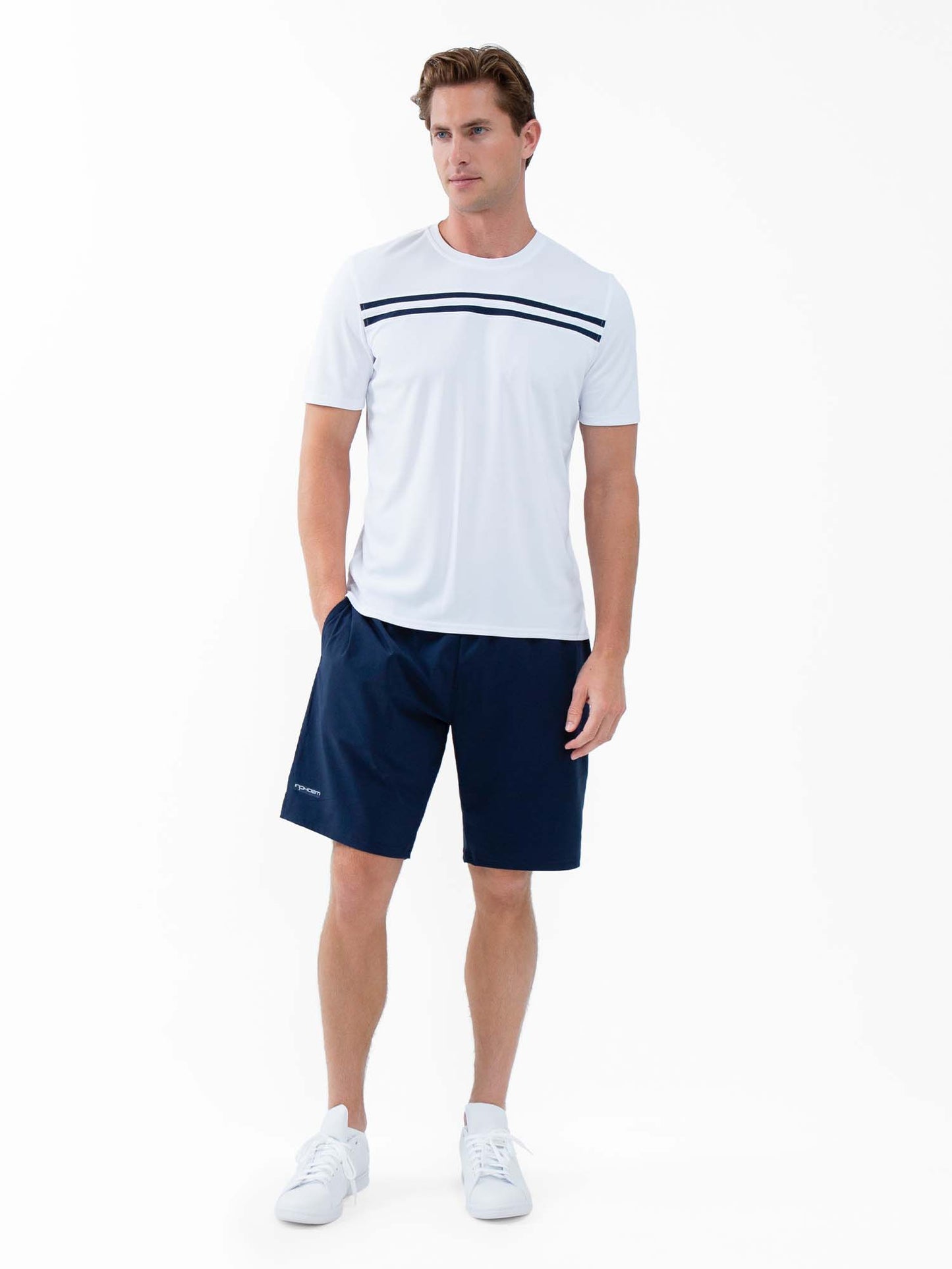 A man with light skin and short brown hair poses against a white background wearing the inPhorm NYC Performance Men's Tournament Short Sleeve Crew Neck in White/Midnight, navy shorts, and white sneakers. His left hand is in his pocket.