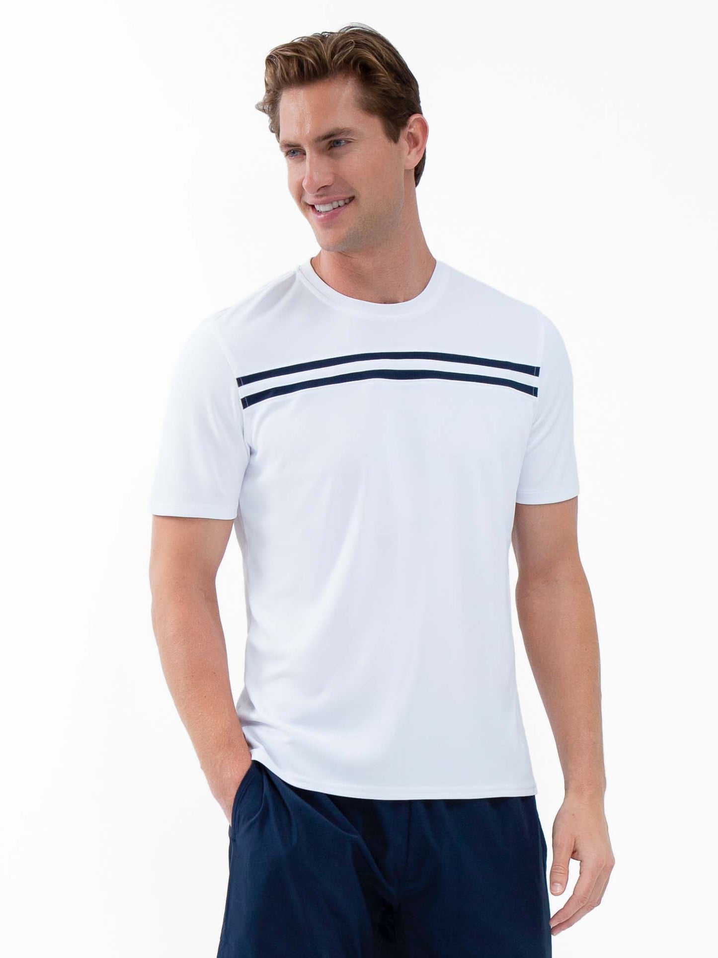 A man wears the inPhorm NYC Performance Men's Tournament Short Sleeve Crew Neck in white/midnight, featuring two thin navy blue stripes. The moisture-wicking shirt keeps him cool as he smiles, hand in his navy shorts pocket against a plain white background.