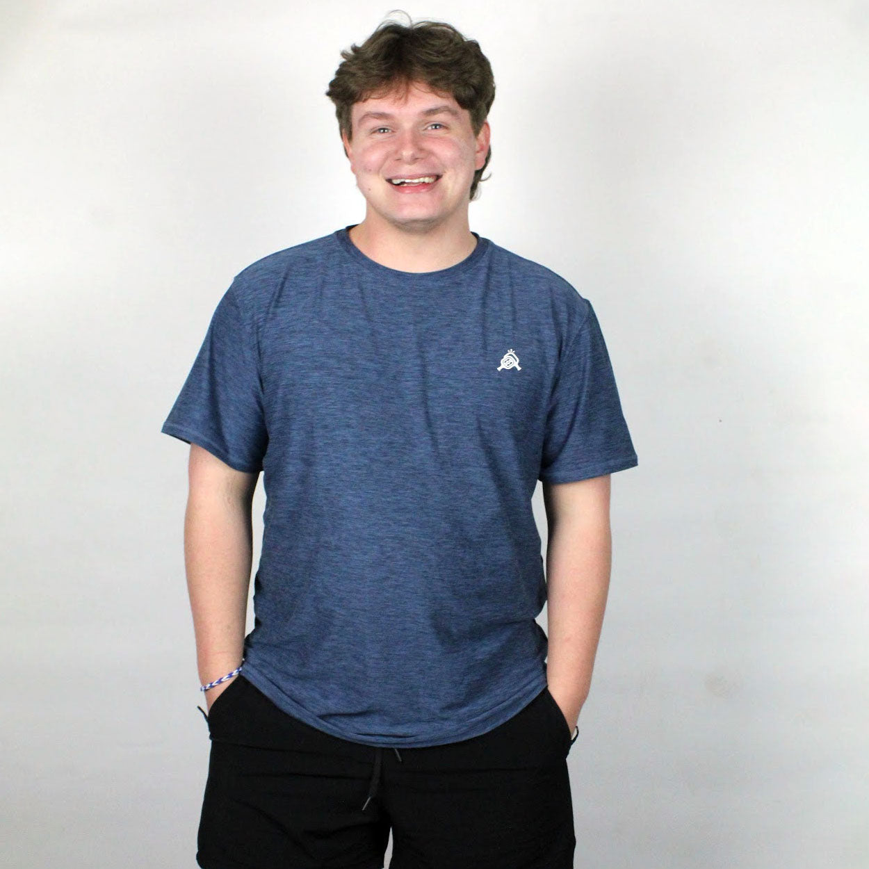 A young man with short brown hair smiles at the camera, wearing a Good Get AeroBliss Performance Short Sleeve T-shirt in blue with a small white chest logo and black shorts, standing against a plain white background.