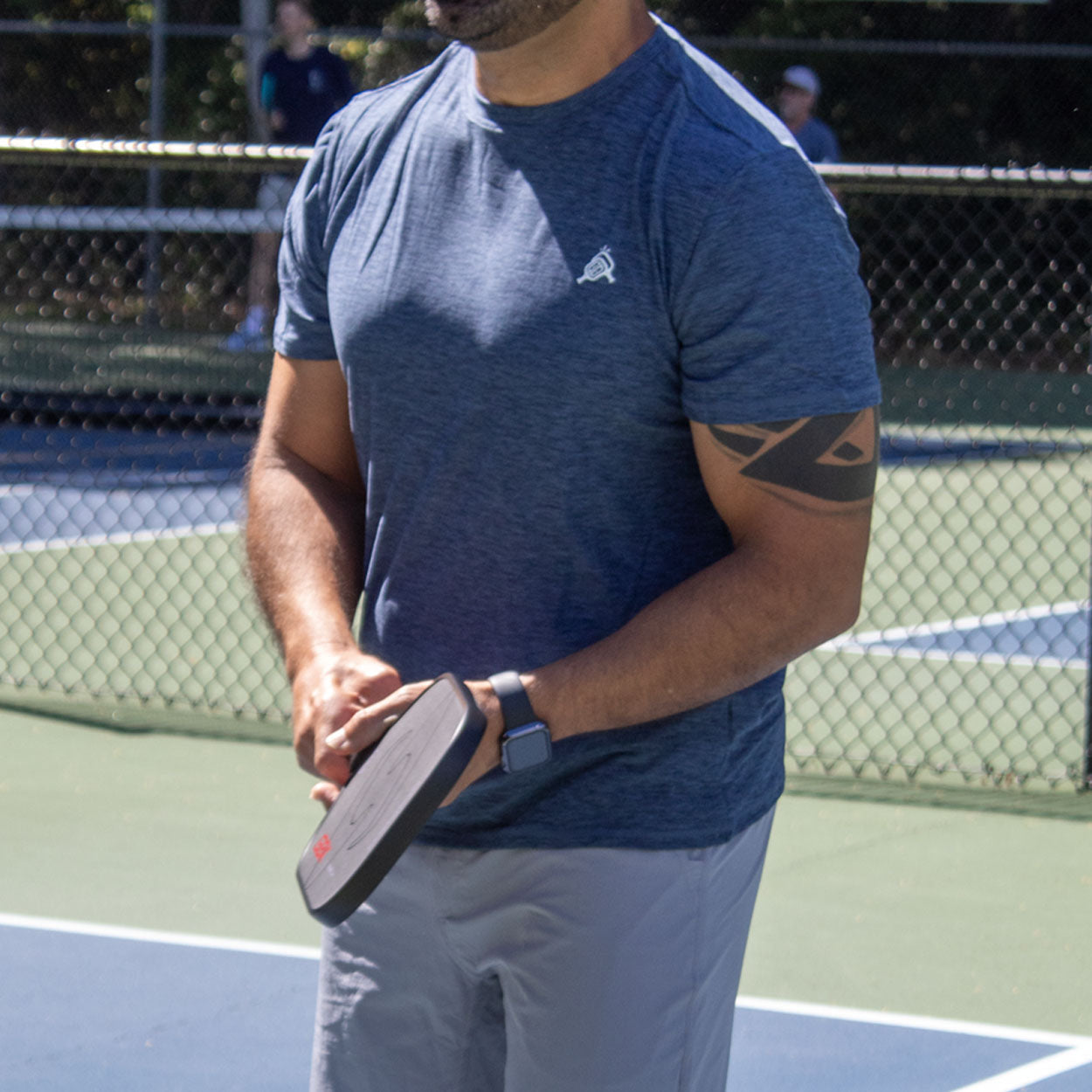 A man wearing the Good Get AeroBliss Performance Short Sleeve T-shirt and gray shorts stands on a tennis court, holding a pickleball paddle. He has a black armband tattoo, a watch on his left wrist, and is near the net.