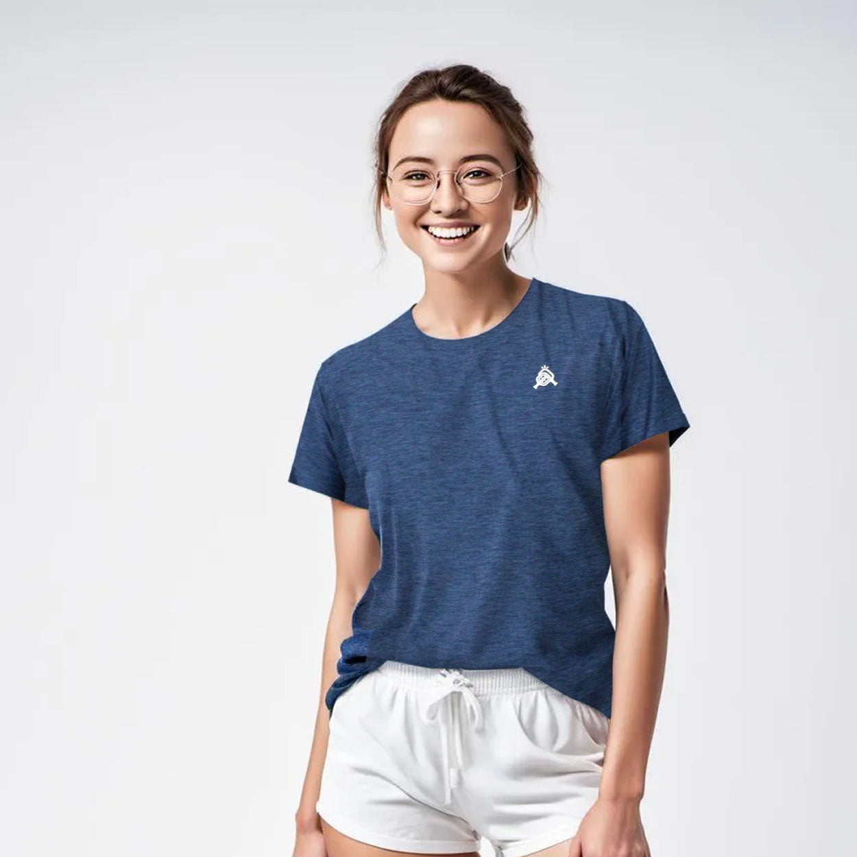 A young woman with glasses and tied-back brown hair smiles at the camera, wearing a blue Good Get AeroBliss Performance Short Sleeve T-shirt with a small white logo on the left chest and white shorts against a plain light background.
