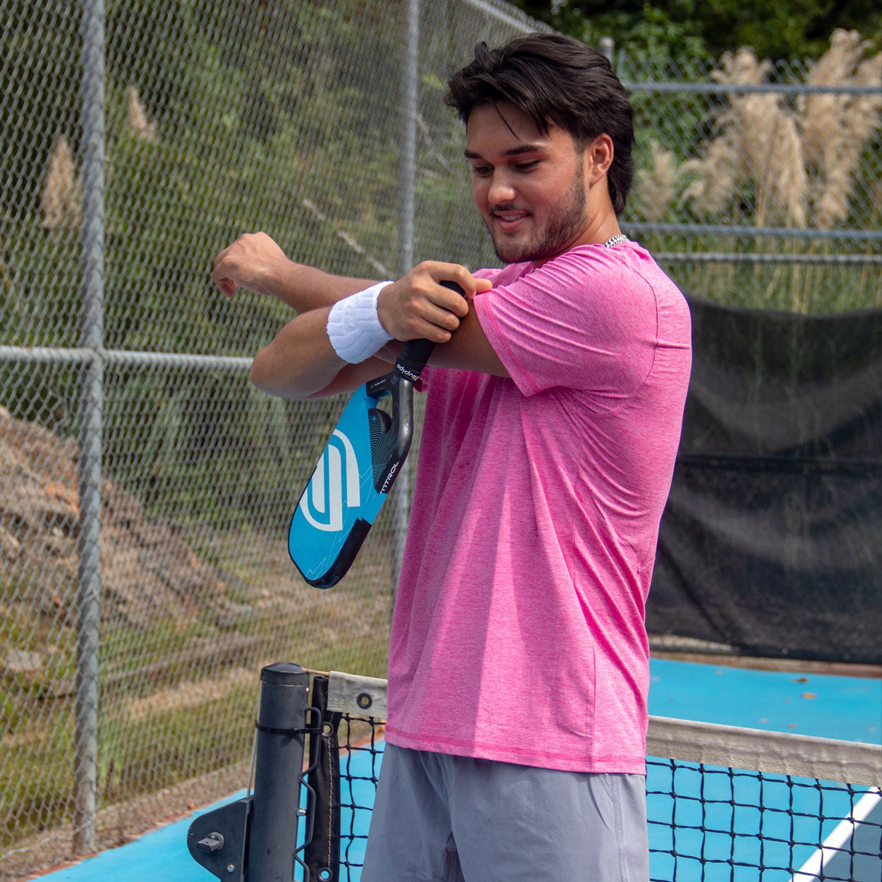 A young man wears the Good Get AeroBliss Performance Short Sleeve T-shirt and gray shorts on a blue pickleball court, pulling up his left sleeve as he holds a paddle. He sports a white wristband by a chain-link fence and black net.