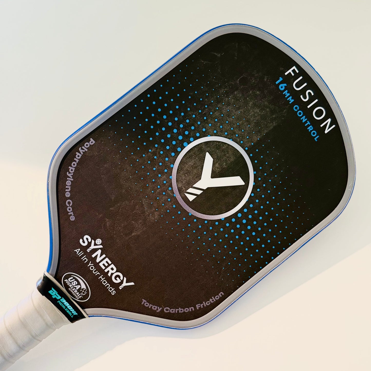 A close-up of a black pickleball paddle labeled FUSION 16 MM CONTROL with blue accents, a white Y logo, and text SYNERGY All in Your Hands and Toray Carbon Friction. Its white grip is ideal for cleaning with the Bodhi Performance PROeraser™.