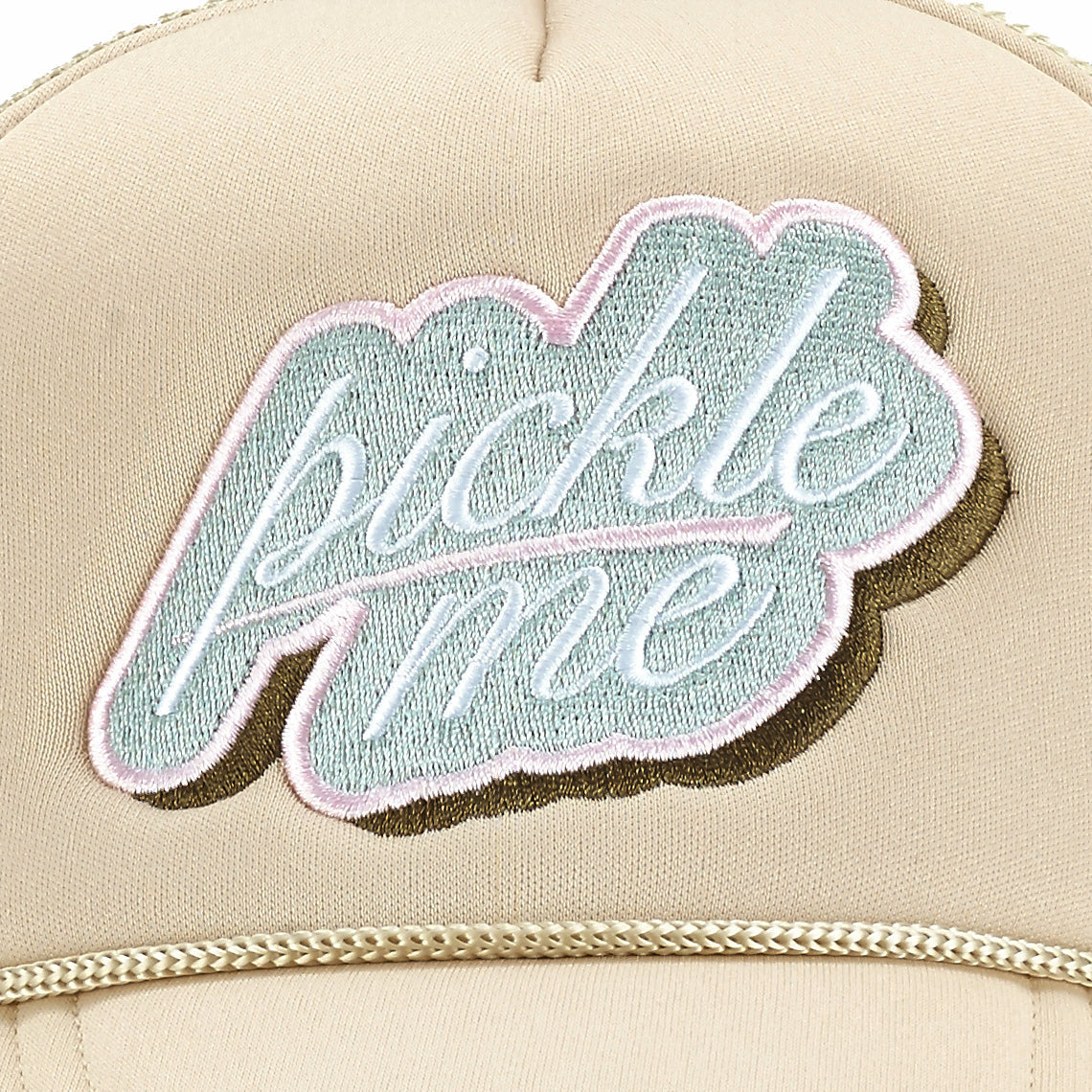 A close-up of a beige ARTI Pickle Me Trucker Hat shows "pickle me" embroidered in light blue with white and brown trim, plus a curved seam and part of the thick, textured brim.