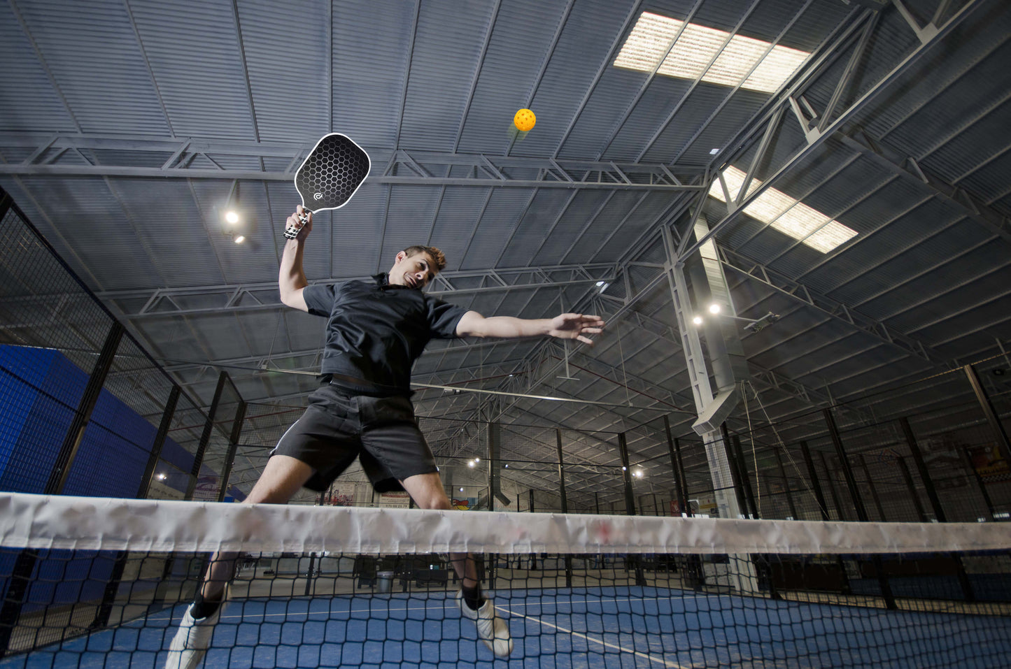 A man in black sportswear leaps on an indoor blue pickleball court, swinging The Big Dink - Model1 Paddle from The Big Dink’s 2-pack Special overhead to hit a yellow ball. Net, fencing, and lights are visible in the dynamic low-angle view.