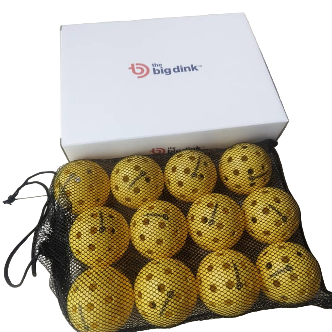 A black mesh bag with twelve yellow The Big Dink - Pickleball Indoor Balls (40-hole design) sits on a tiled floor before a closed white box labeled The Big Dink. The balls are neatly arranged, ready for your next game.