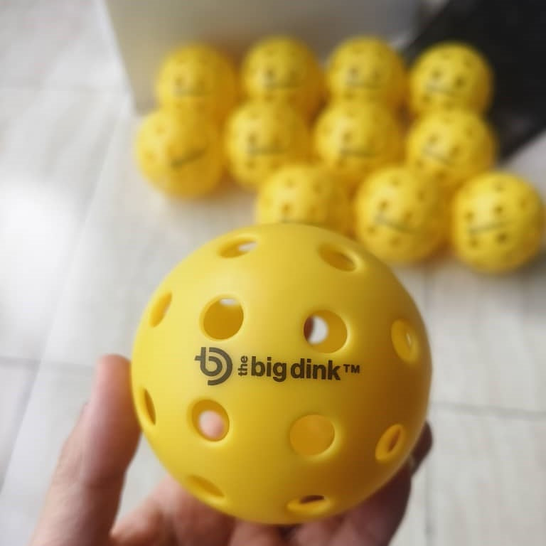 A hand holds a yellow The Big Dink - Pickleball Indoor Ball by The Big Dink, with several identical balls from the 12 Pack scattered across a light-colored tiled floor in the background.