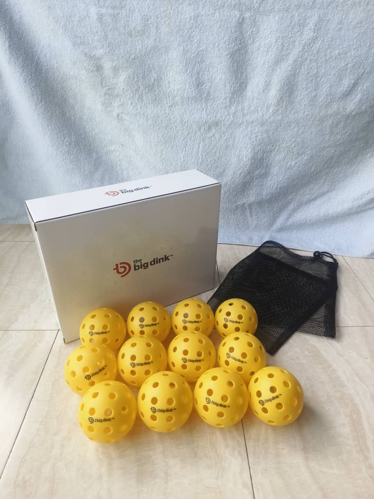 Twelve yellow The Big Dink - Pickleball Indoor Balls from The Big Dink are arranged in front of a white box, with a black mesh bag beside them on a tiled floor and light blue fabric in the background.