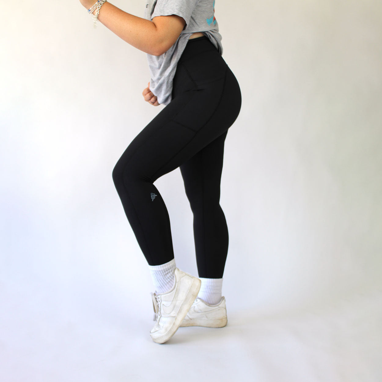 A person wears Good Get Dinks & Clinks Women's Pickleball Leggings with a phone pocket, white crew socks, white sneakers, a loose gray t-shirt, and bracelets on one arm. They stand sideways on a light background with their head out of frame.
