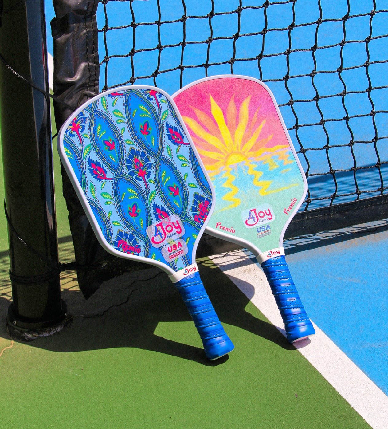 Two Damask 4Joy Pickleball Paddle Premio Series paddles by 4Joy Paddles rest against a net on a green and blue outdoor court, showcasing vibrant designs—a blue peacock feather print and a sun motif—with blue handles and thermoformed edges.