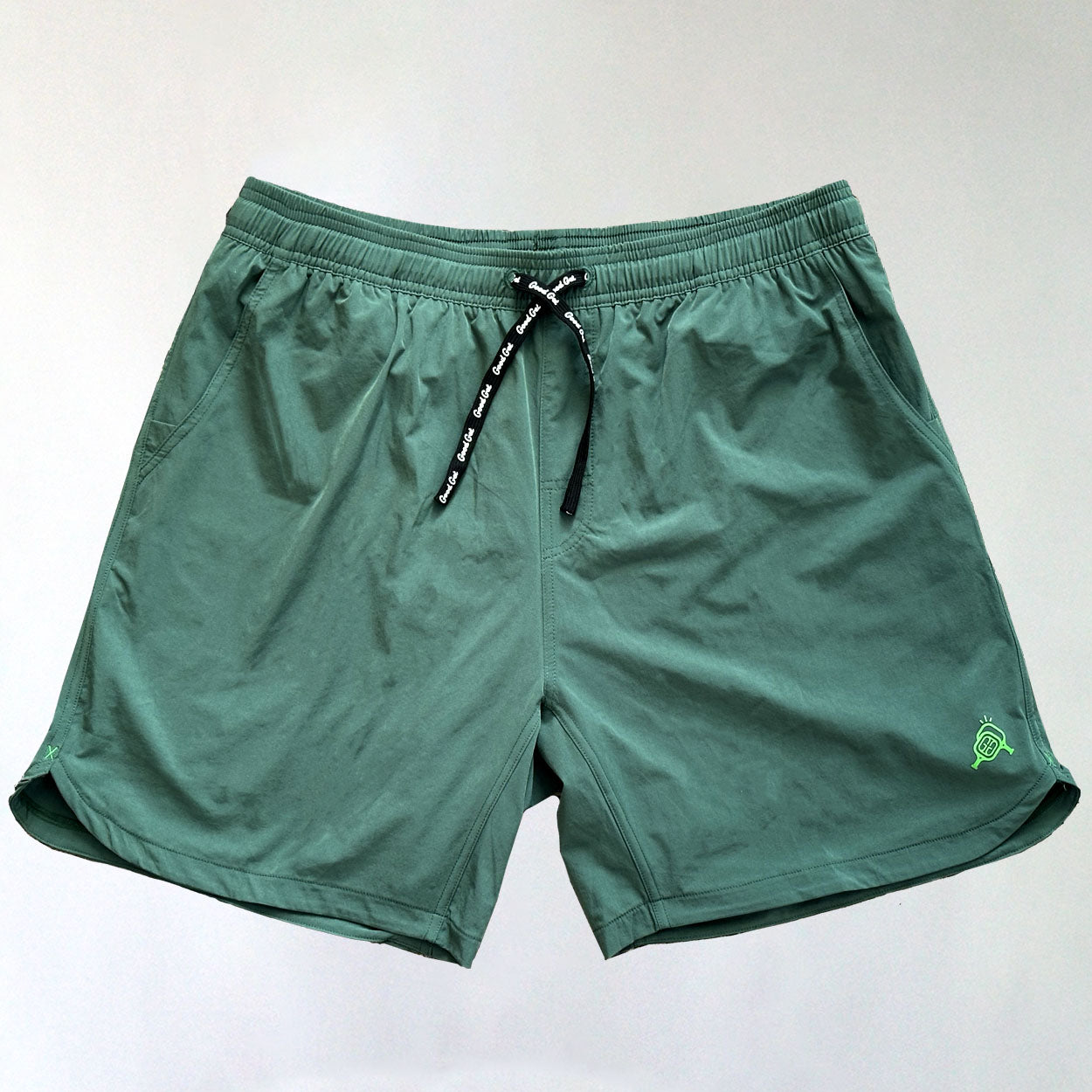 A pair of Good Get All Eyes On Me Pickleball Shorts in green, featuring an elastic waistband, black drawstring with white text, side pockets, 6-way stretch fabric, and a small light green stitched logo on the lower left leg.
