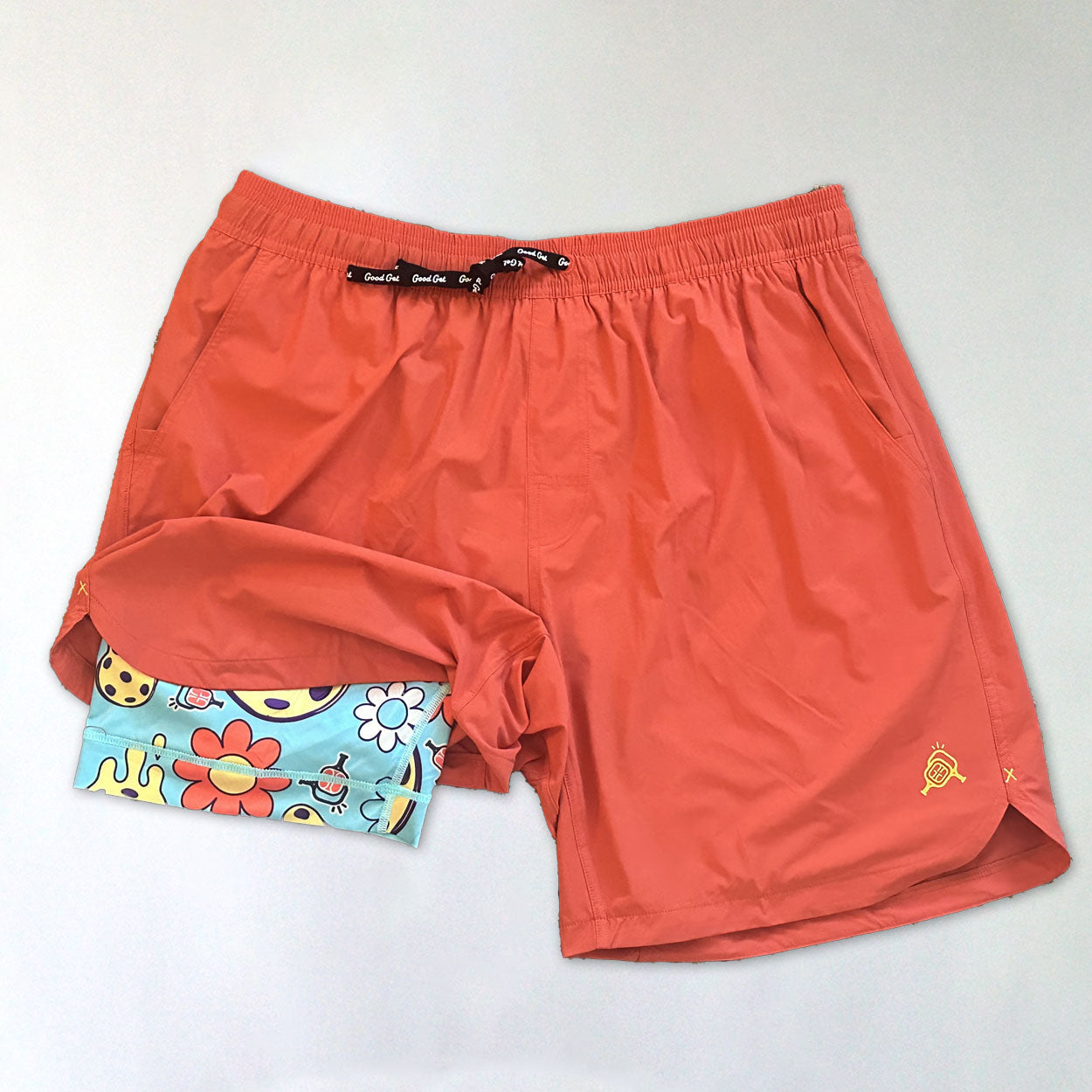 A pair of coral-orange swim trunks with a black drawstring and a small yellow logo on the left leg, partially tucked inside folded Good Get Pickleball Shorts, Drippy Edition, featuring 6-way stretch and a playful floral and smiley face pattern.
