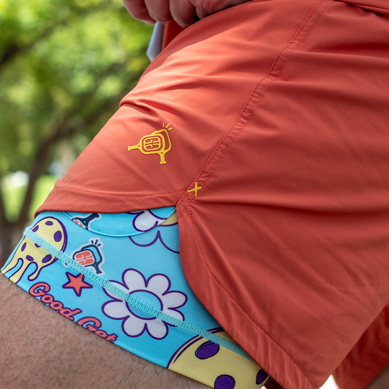 A close-up of a leg wearing Good Get Pickleball Shorts, Drippy Edition, in orange with a yellow beaker logo over blue, 6-way stretch compression shorts featuring cartoon cats, flowers, and "Good Cat" text. Blurred trees appear in the background.