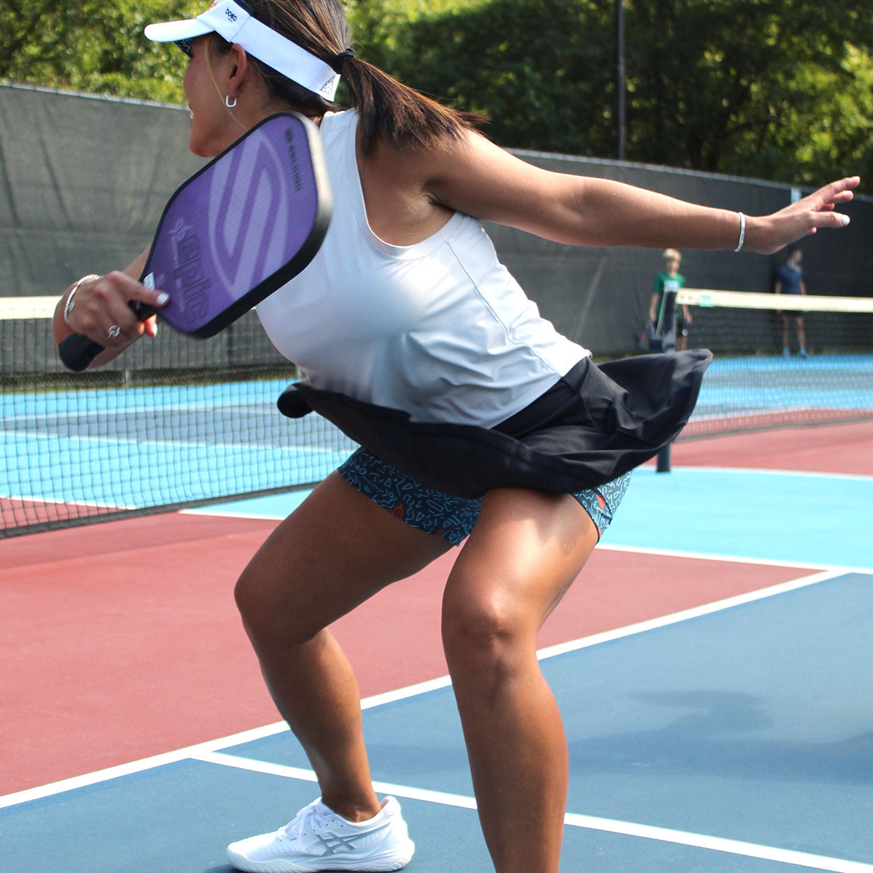 On a sunlit blue and red court, a woman in athletic wear crouches and swings a paddle, sporting the Good Get Skirtin' Round the Kitchen – "Court Clash Edition" skirt with compression liner, plus a white tank top, visor, and sneakers.