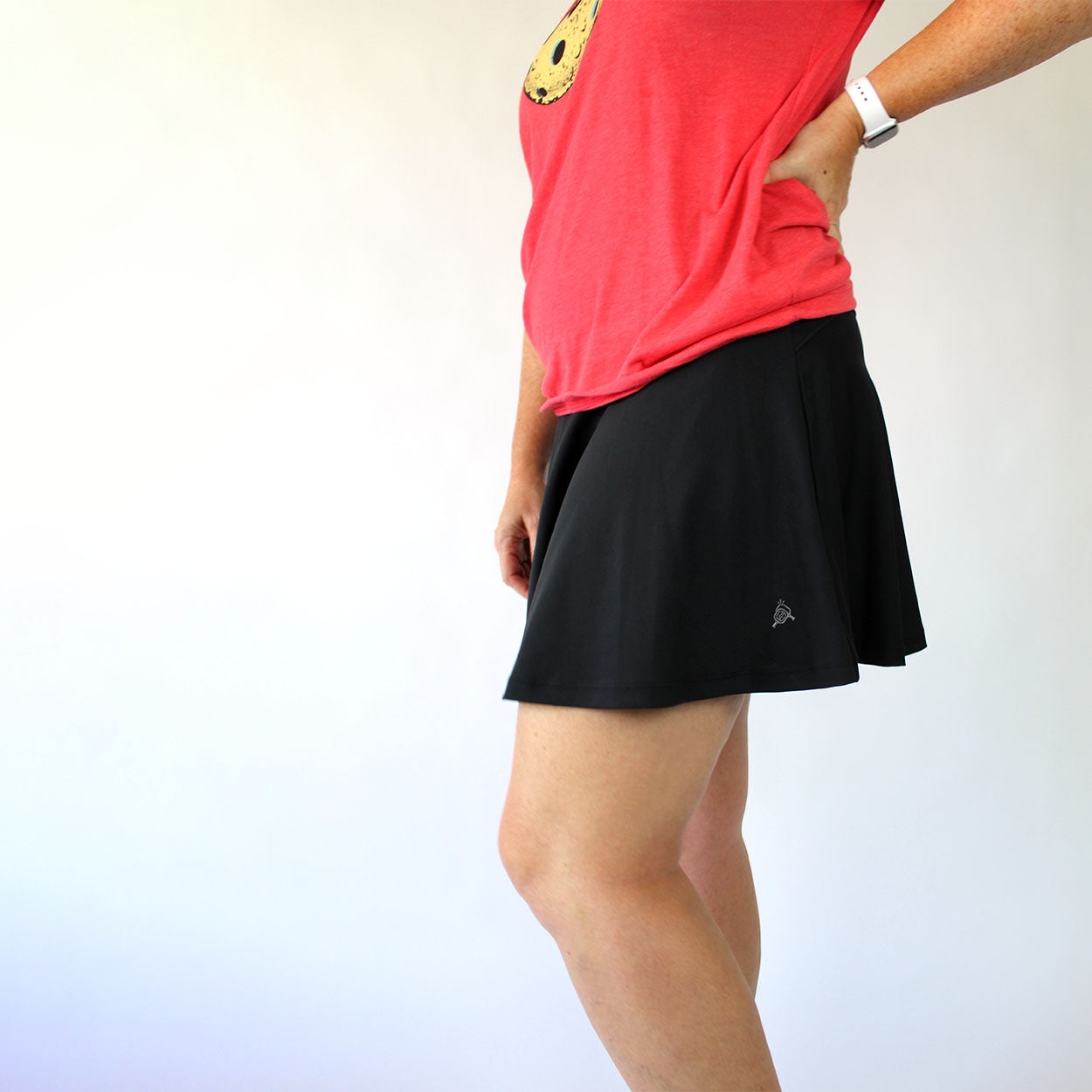 A person stands sideways against a plain background, wearing the Good Get Skirtin' Round the Kitchen - "GG Edition" Pickleball Skirt. Their left hand rests on their hip with a white smartwatch; their face isn’t visible.