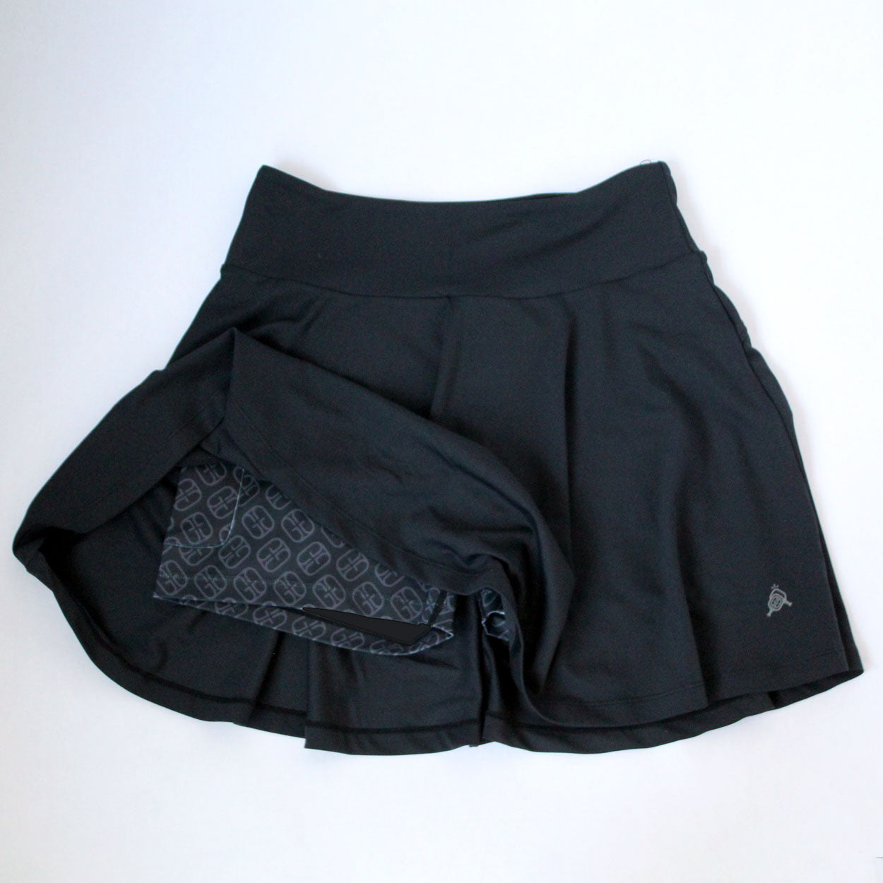 The Good Get Skirtin' Round the Kitchen "GG Edition" pickleball skirt is black with a flared outer layer and geometric moisture-wicking inner shorts, shown flat on a white surface with one side lifted to display the shorts.