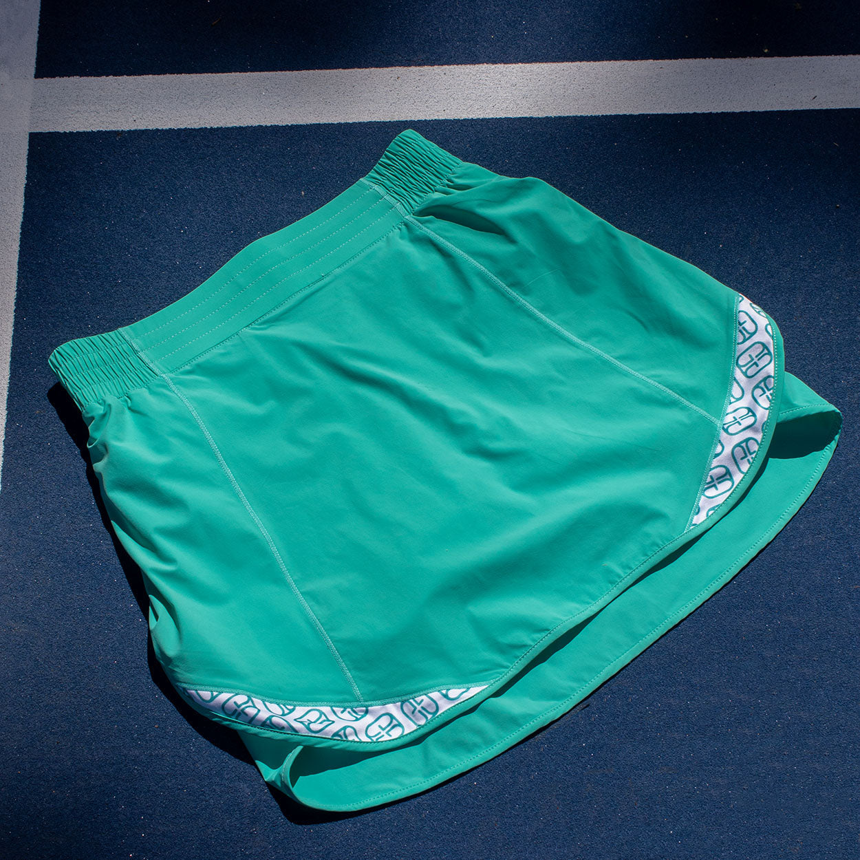 The Good Get Lob Deep Pickleball Skirt, featuring bright teal fabric, a wide elastic waistband, and white patterned trim, rests on a blue court in sunlight and shadow—moisture-wicking and designed for active play near the white line.