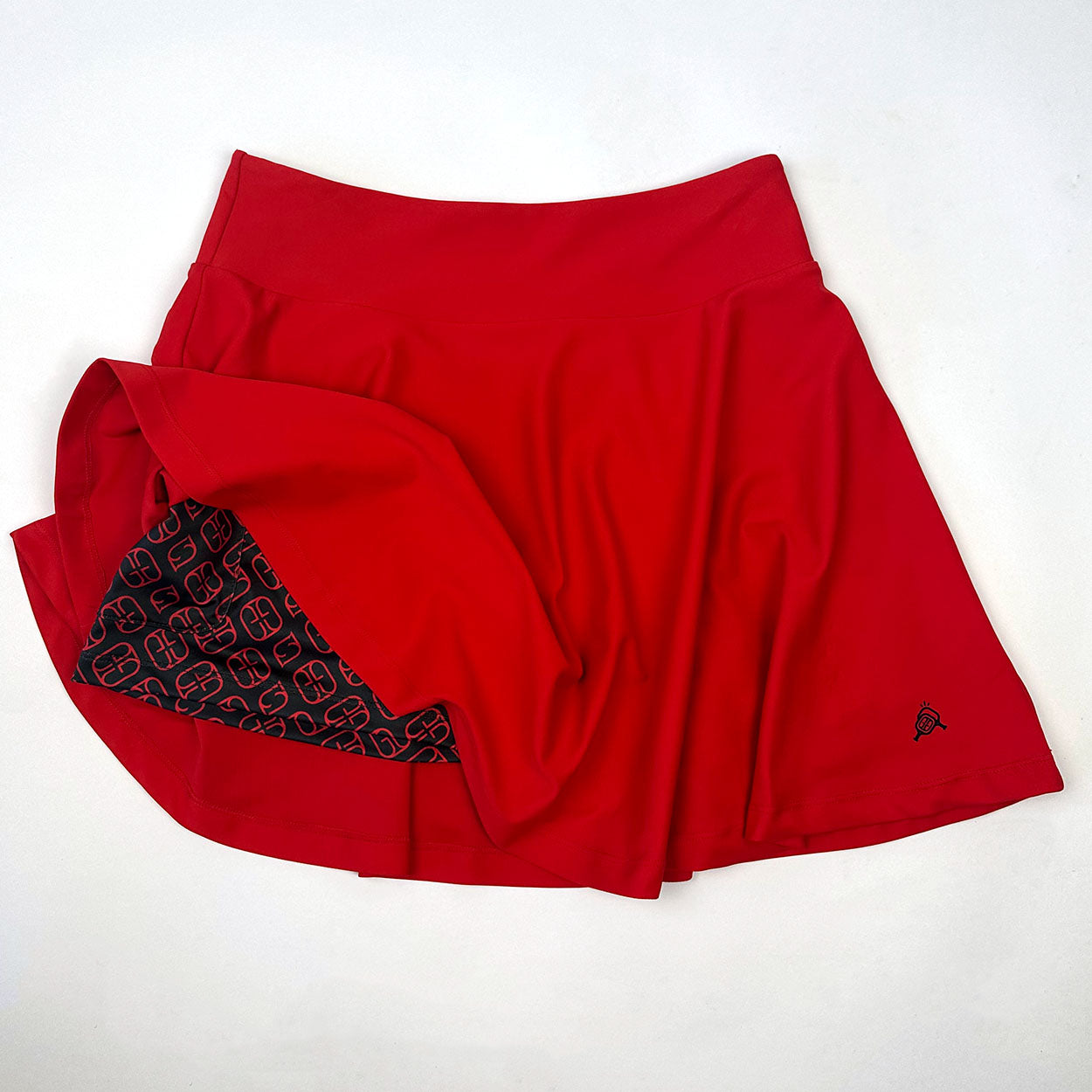 The Good Get Skirtin' Round the Kitchen - Rally Red Edition pickleball skirt is laid flat on a white background, partially lifted to show dark geometric compression shorts and features a small embroidered black logo near the right hem.