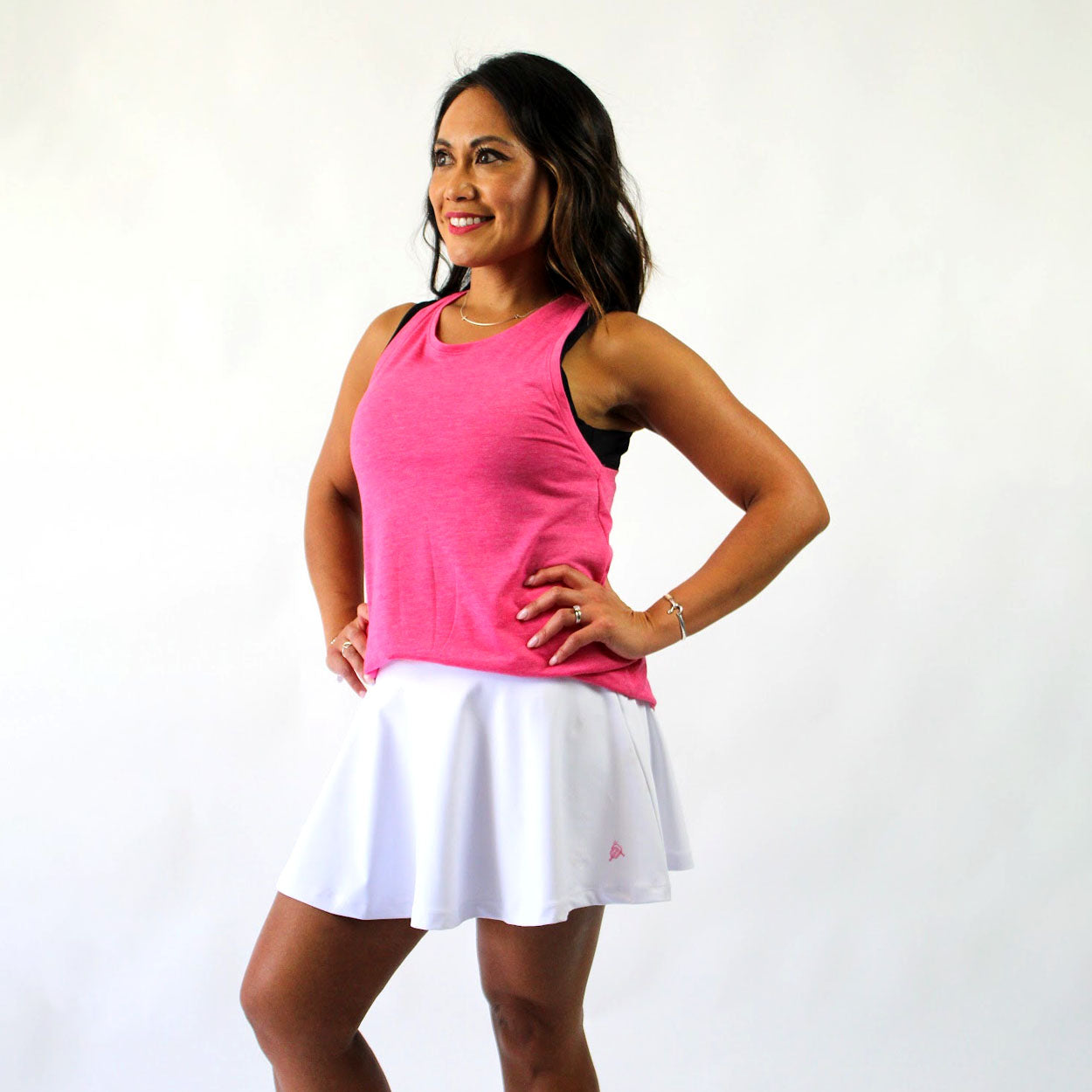 A woman with long dark hair smiles confidently, wearing Good Get's Skirtin' Round the Kitchen - "You're A Star Edition" Skirt, a white athletic skort with built-in compression shorts, paired with a pink sleeveless top on a plain white background.