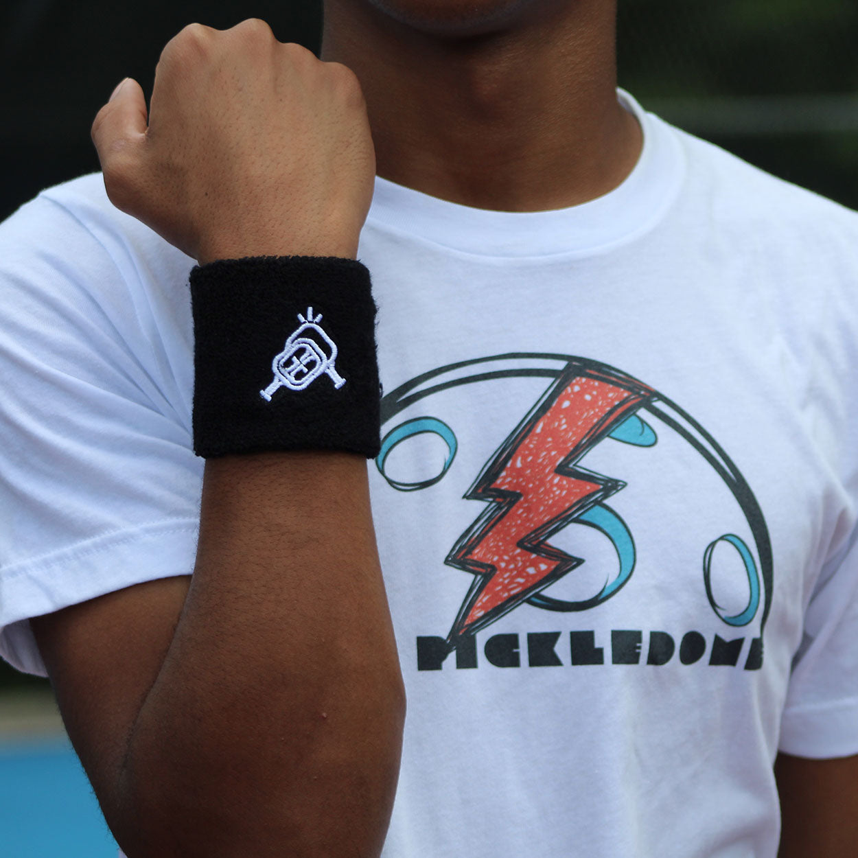 A person wearing a white graphic t-shirt with a red lightning bolt and circles, plus a black Good Get Wristband of Dinking +1 featuring a white embroidered barbell with halo. Their left arm is raised, background blurred.