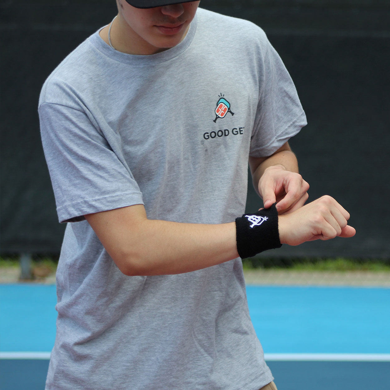 A person in a light gray Good Get t-shirt and black cap stands on a blue tennis court, adjusting the Wristband of Dinking +1 by Good Get on their left wrist. Their head is partly out of frame, with a blurred background and black fence.