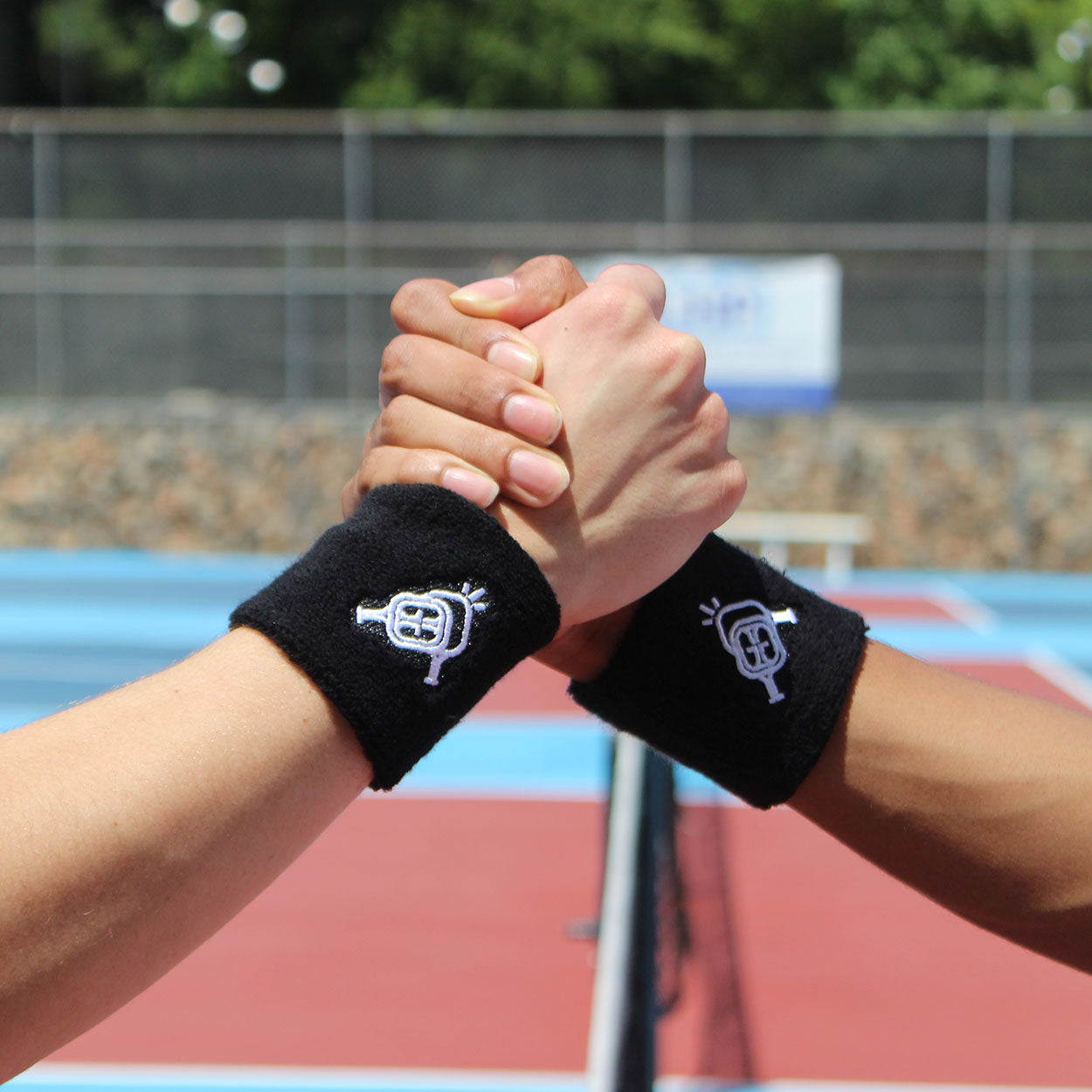 Two people clasp hands over a tennis net on an outdoor court, each wearing the Good Get Wristband of Dinking +1 with a white embroidered logo. In the background are a fence, greenery, and a blurred sign.