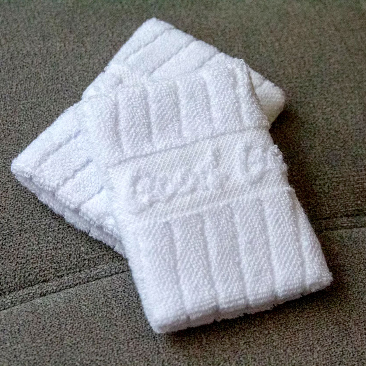 Two folded Good Get Wristband of Dinking +1 pickleball wristbands with ribbed white terry cloth and a subtle horizontal stripe featuring embossed text rest on a gray, textured fabric background.