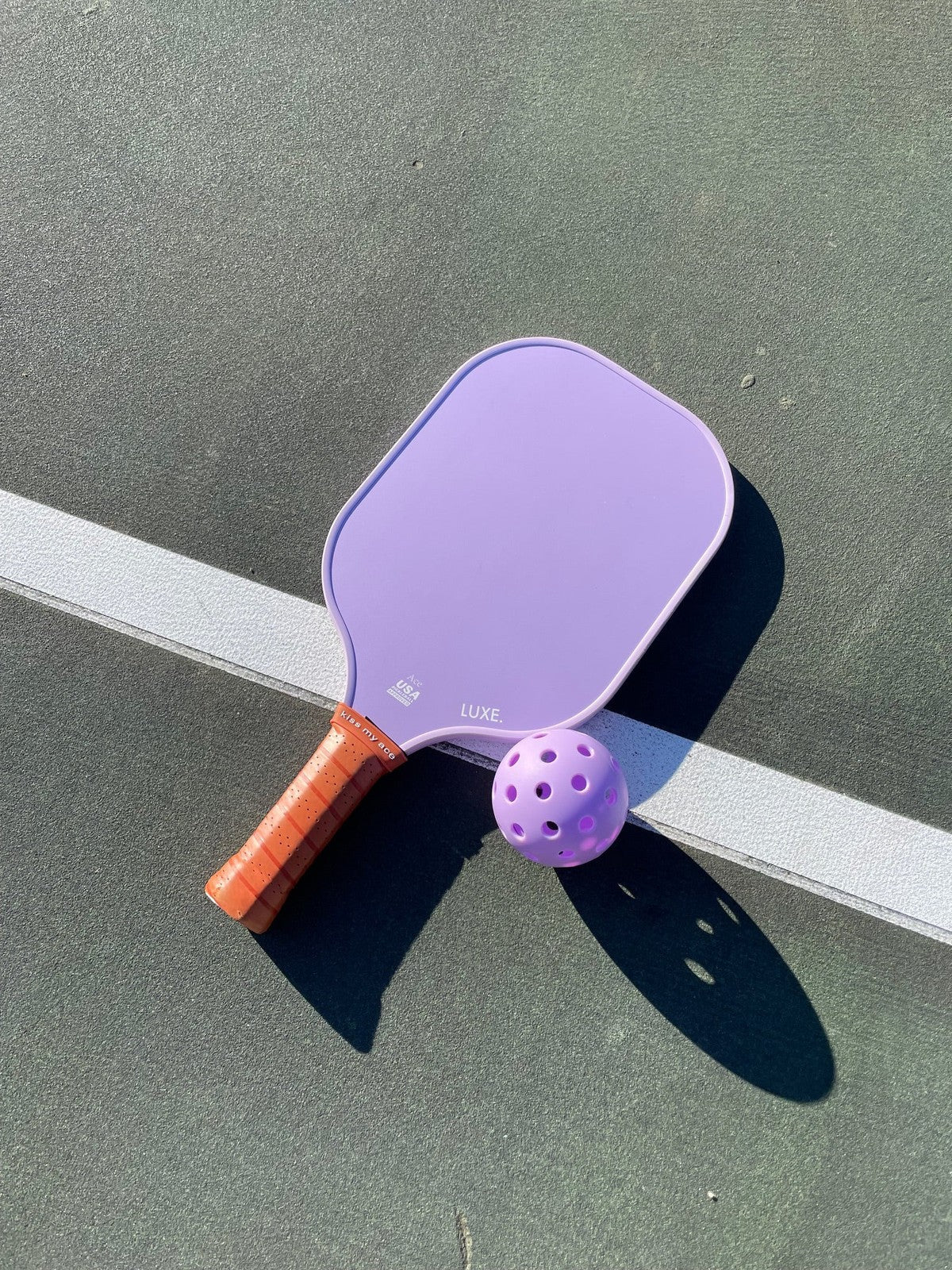 A purple paddle with a brown plaid handle and a LUXE Pickleballs high-visibility purple ball by LUXE Pickleball are on a green court near a white boundary line, with shadows cast to the right.