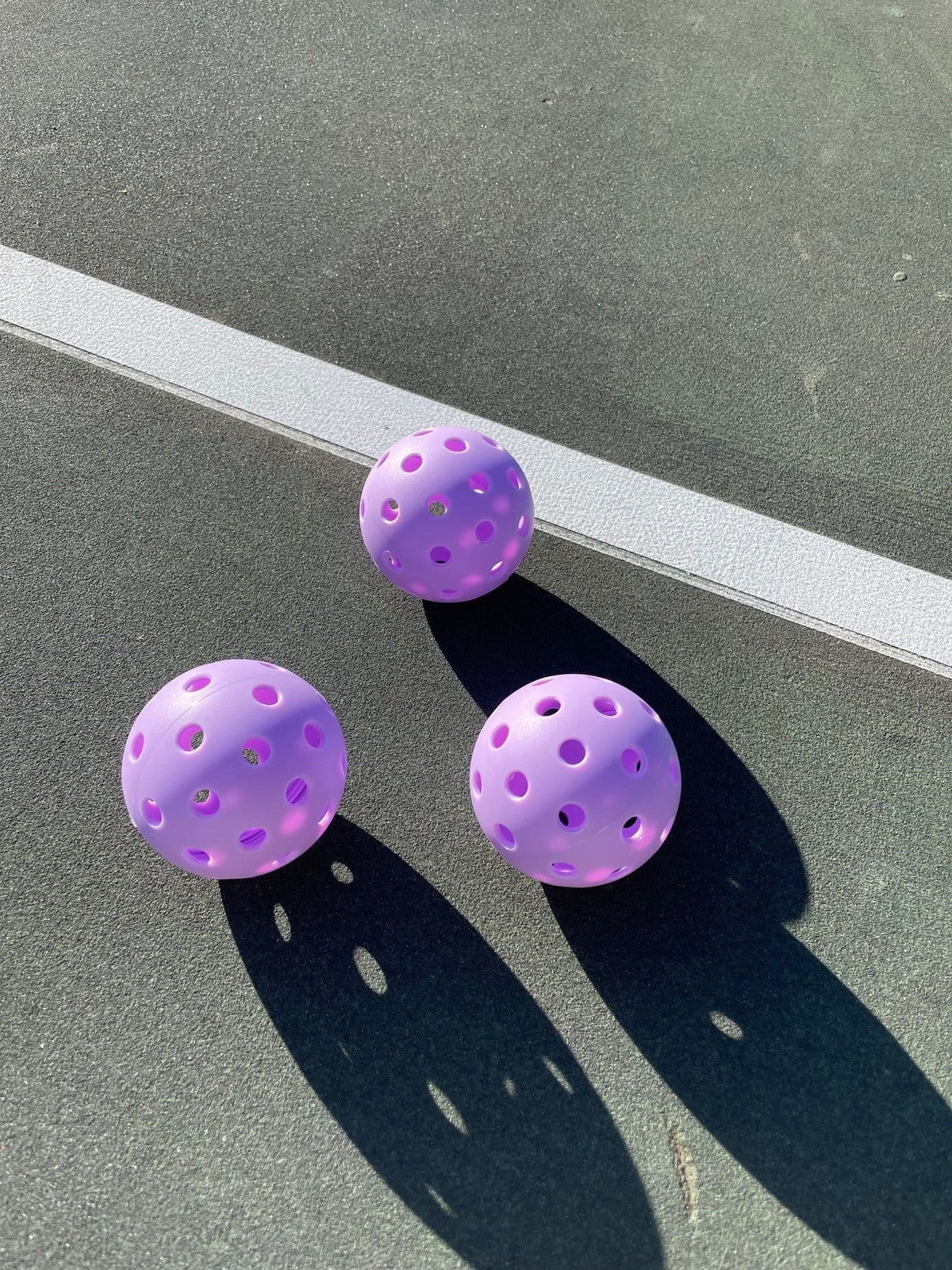Three purple LUXE Pickleballs by LUXE Pickleball rest on a sunlit green court, casting shadows. Two durable balls are near the white boundary line while one sits farther above it, their high visibility ideal for outdoor play.