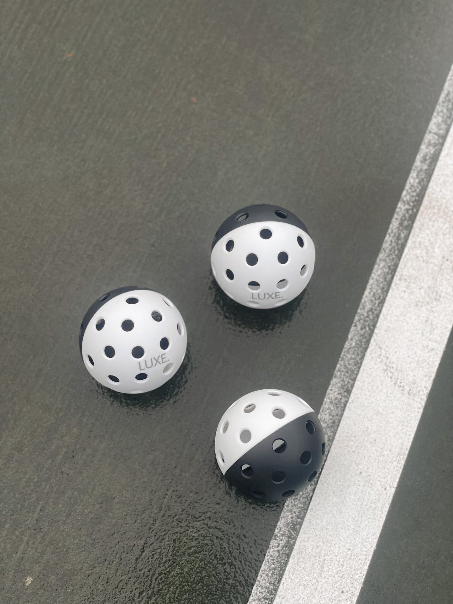 Three black-and-white LUXE Pickleballs from LUXE Pickleball, known for their durability in indoor and outdoor play, rest on a wet, dark-green court near a white line. The slightly shiny surface highlights the quality tournament balls.