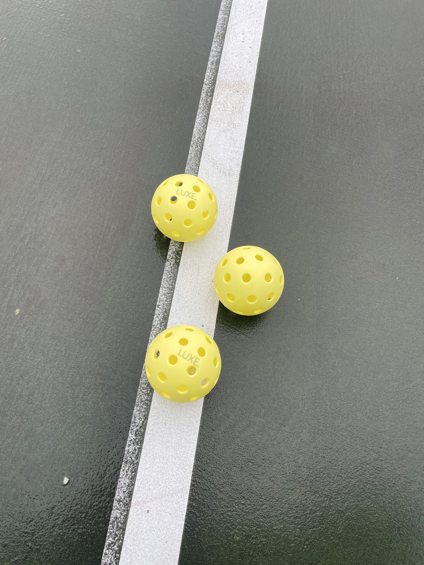 Three yellow LUXE Pickleballs by LUXE Pickleball lie on a dark green court beside a white boundary line. Their textured surface showcases these durable practice and tournament balls for indoor and outdoor play.