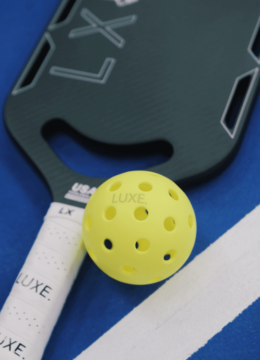 A bright yellow LUXE Pickleballs - Durable Practice and Tournament Ball by LUXE Pickleball sits on a black paddle with geometric lines on a blue court, perfect for indoor or outdoor play.
