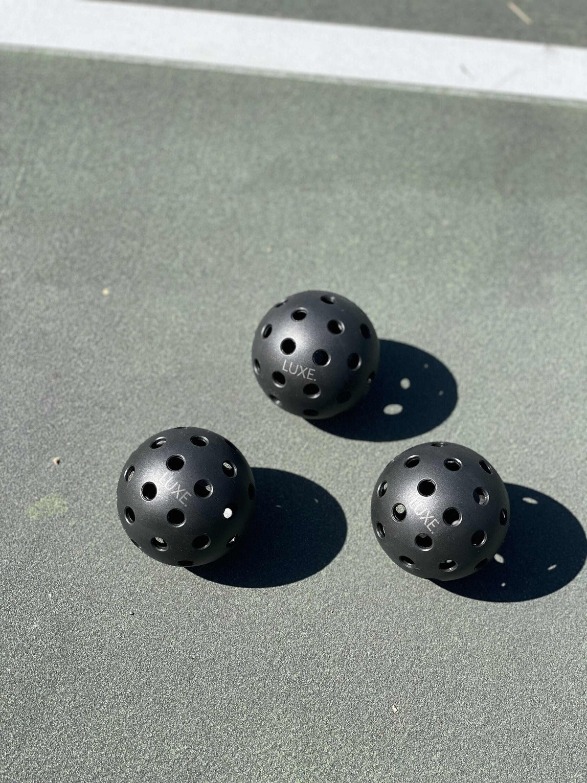 Three LUXE Pickleball practice and tournament balls from LUXE Pickleball rest on a sunlit outdoor court. The black, durable balls feature evenly spaced holes and "LUXE" branding, casting sharp shadows on the high-visibility surface.
